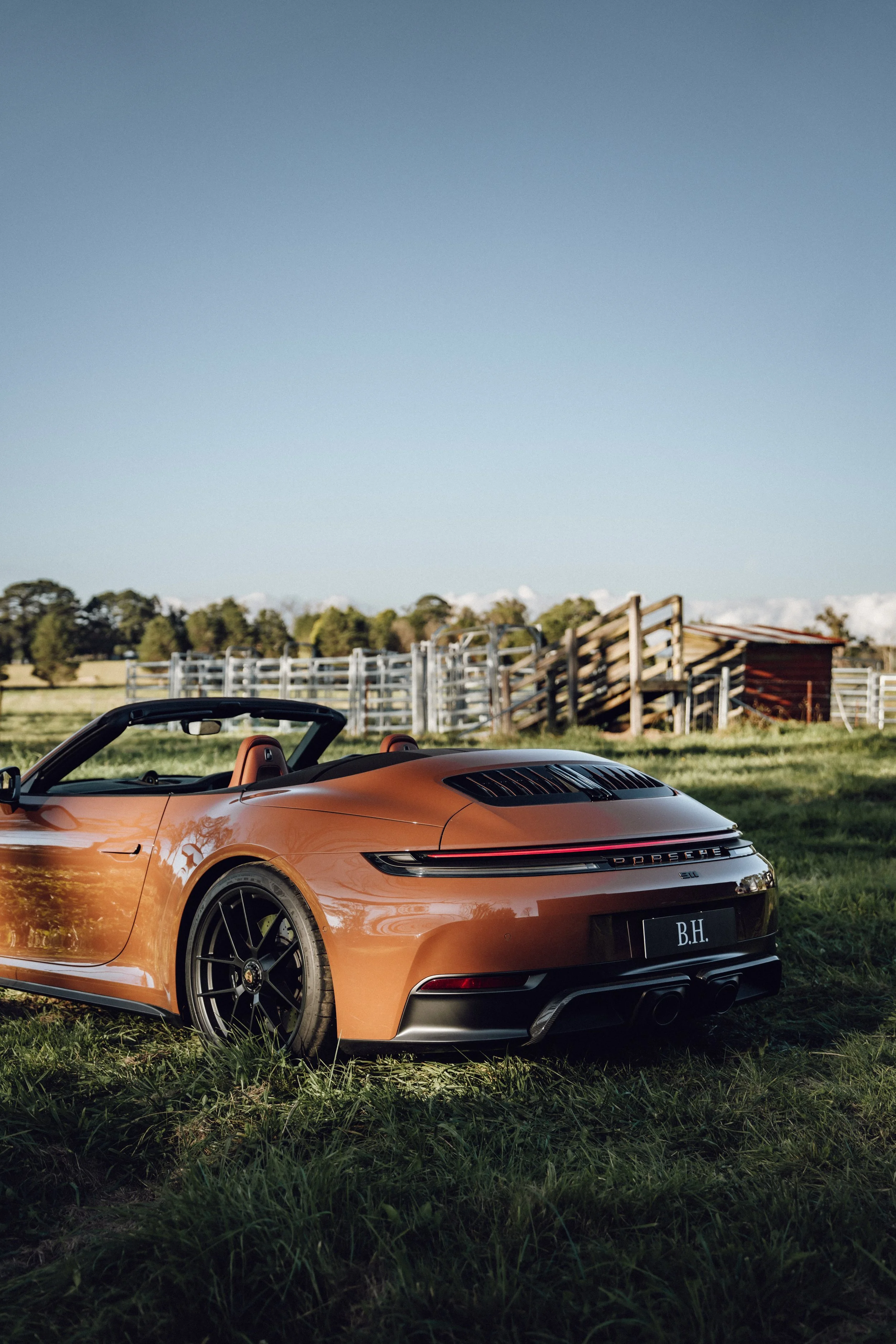 Orange Porsche 911 convertible sports car parked on grassy field with a wooden fence and small barn in the background, under clear blue sky.