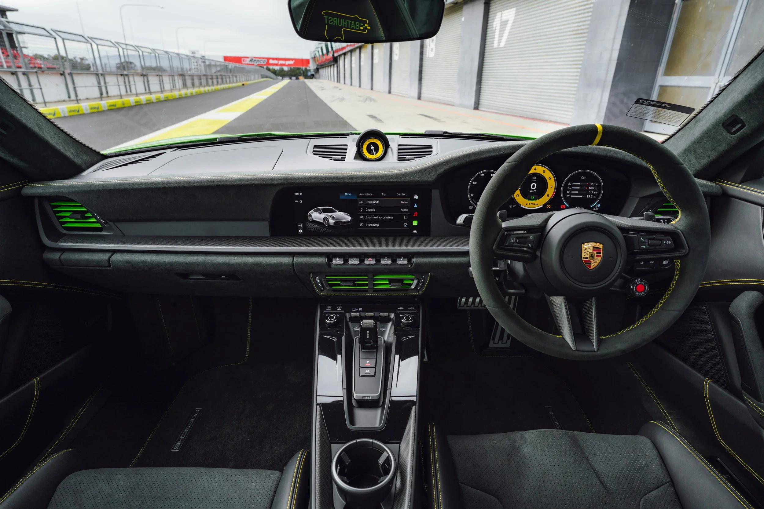 Interior view of a Porsche race car dashboard and steering wheel parked on a racing circuit with closed garage doors on the right and a race track ahead.