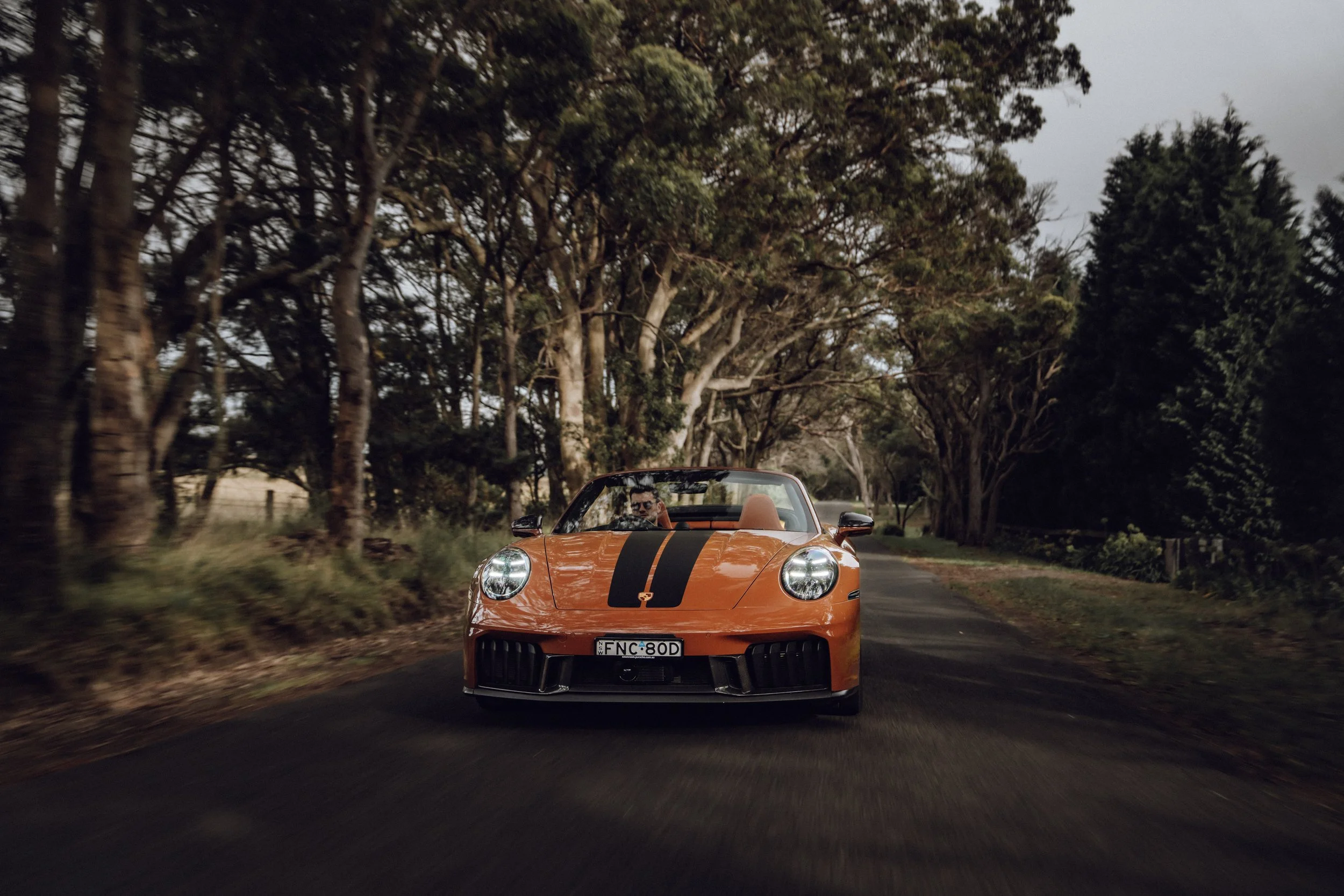 An orange sports car with black racing stripes driving on a rural road surrounded by trees.