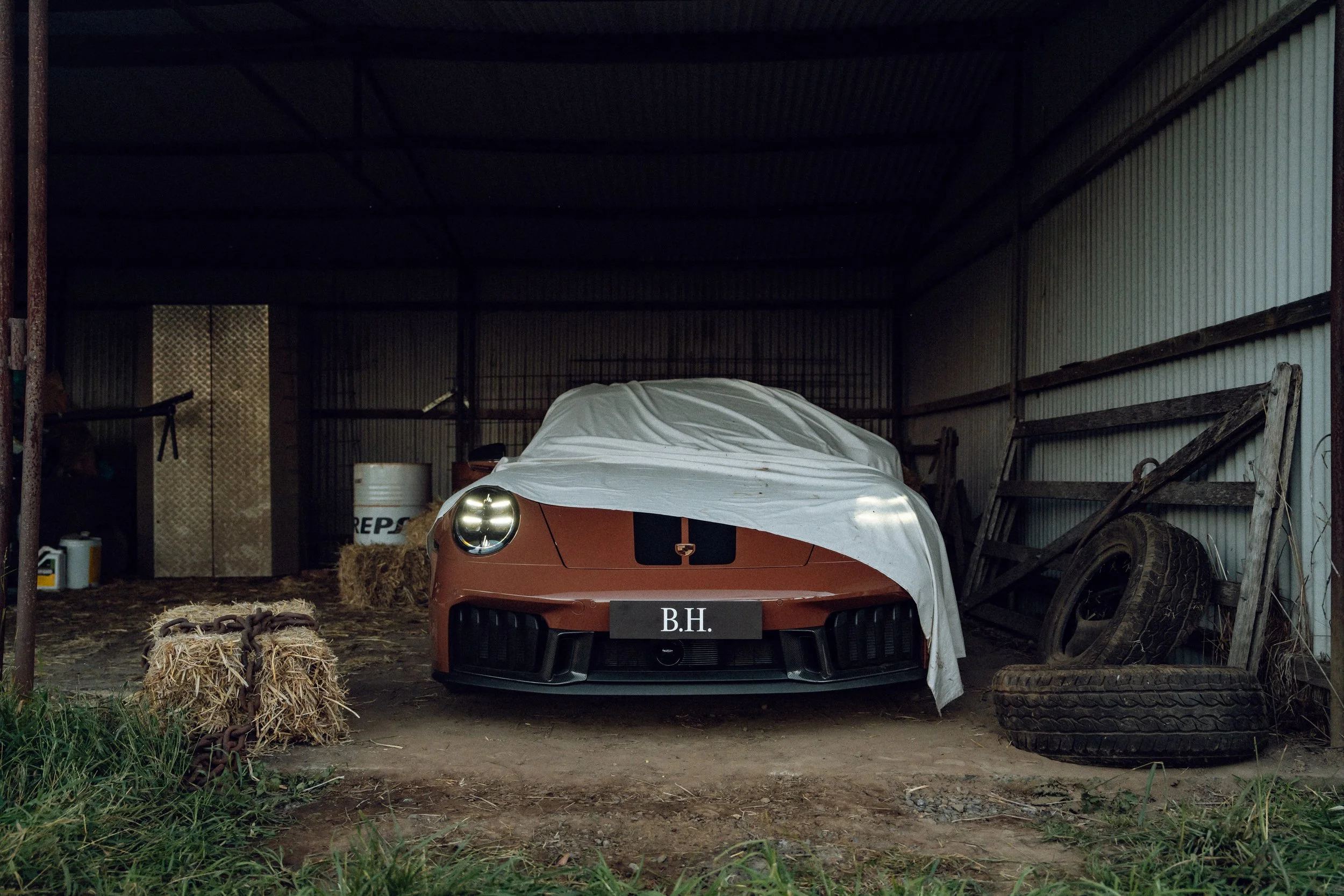 A vintage brick-colored Porsche 992 GTS Cabriolet covered with a white sheet, parked inside a rustic garage with tires and old machinery around.