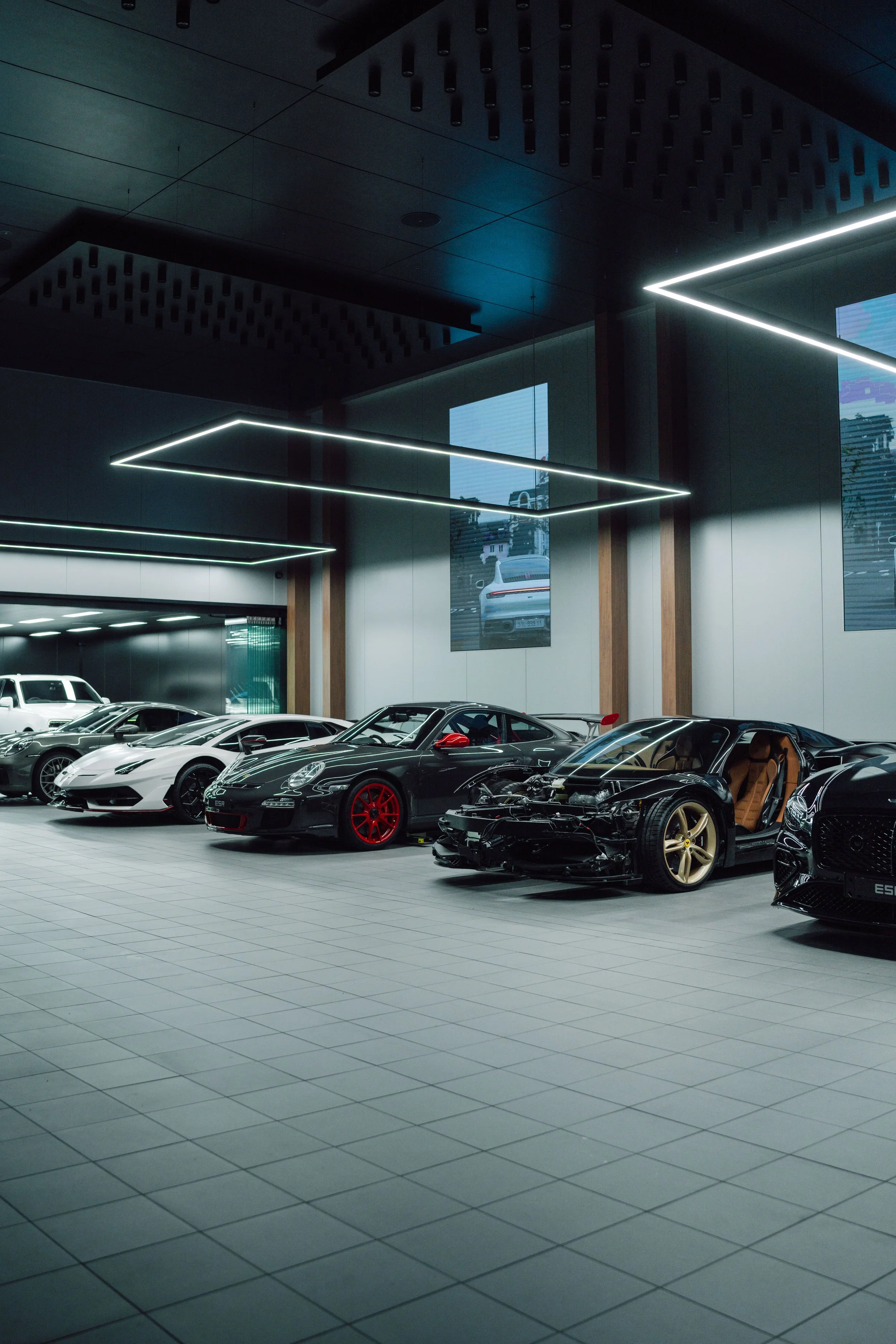 Luxury cars, including a black sports car with an open hood, are displayed in a modern showroom with sleek lighting and large windows.