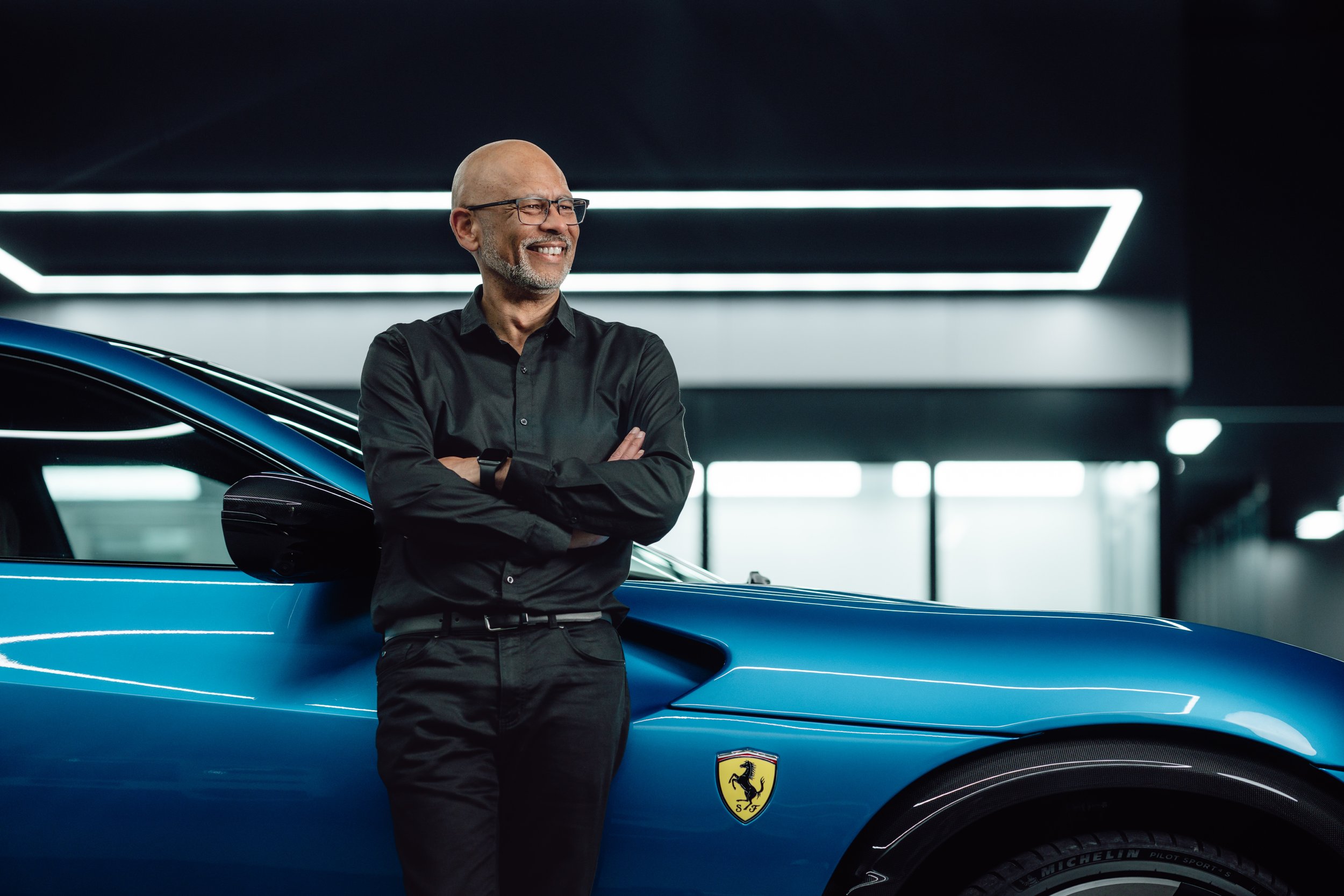 A man with glasses and a beard, wearing a black shirt, standing with arms crossed next to a blue Ferrari sports car in a modern, well-lit showroom.