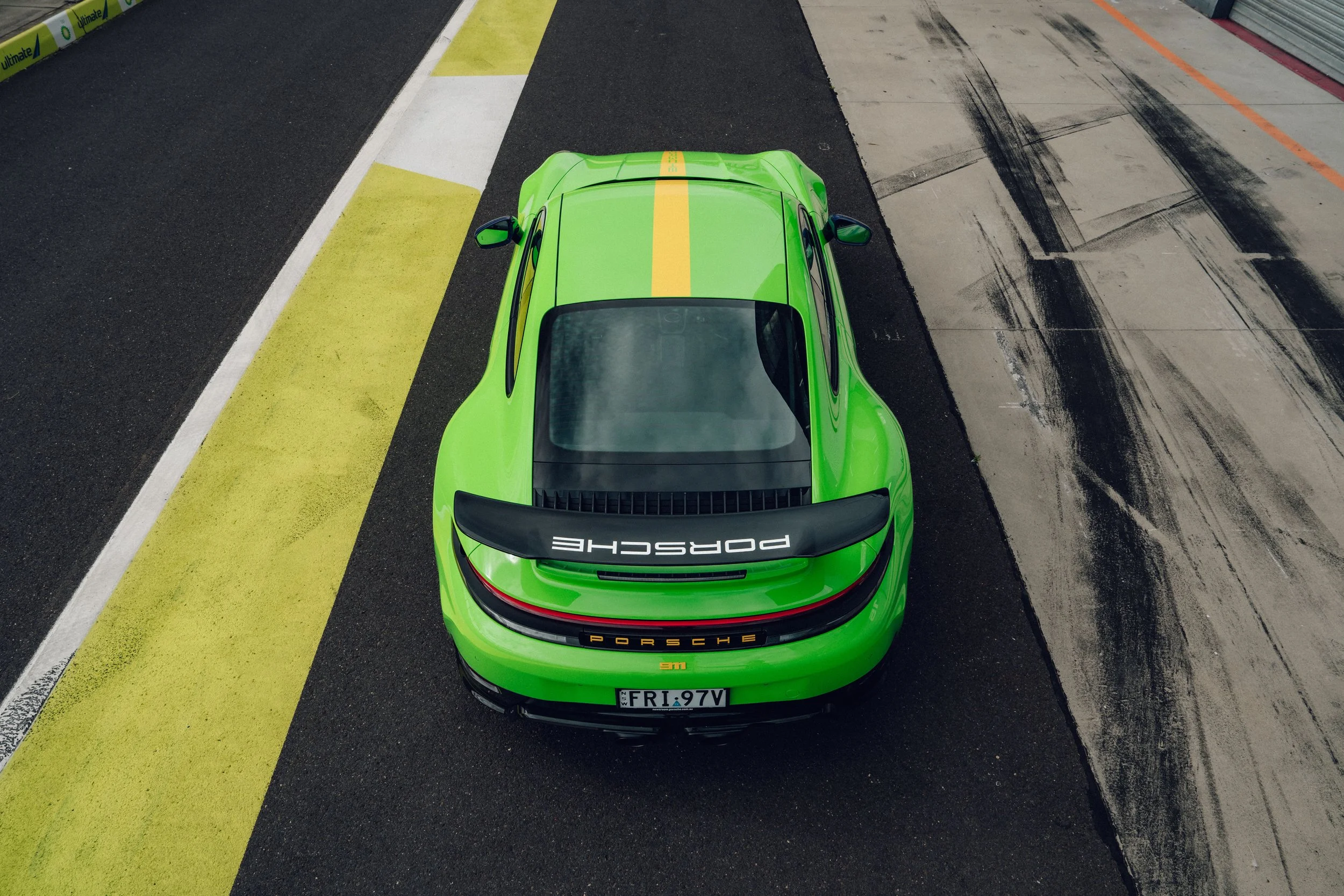 Green Porsche sports car viewed from above, parked on a race track with yellow and white curb on its left and black and gray run-off area on its right.