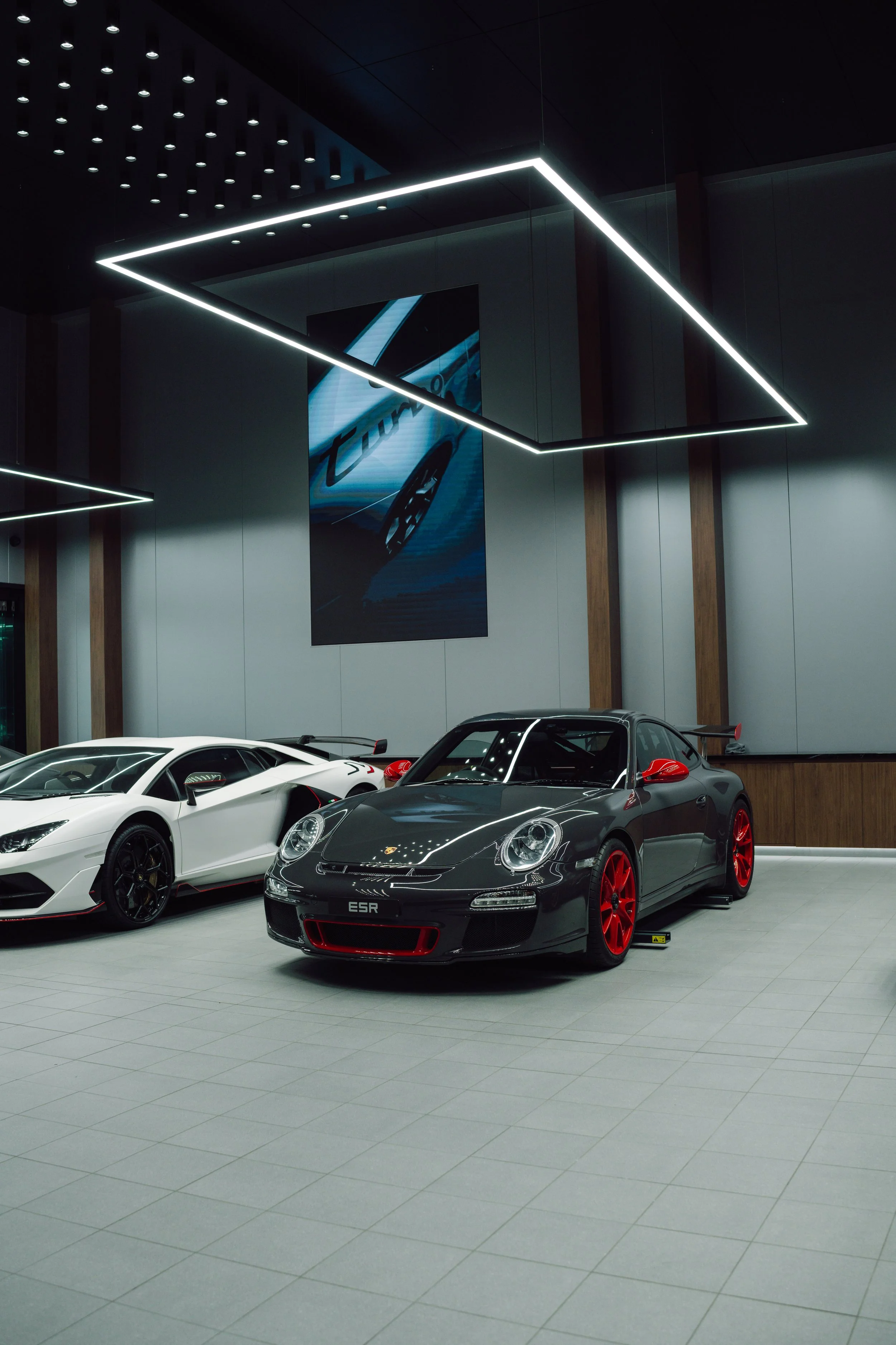 Inside a car showroom with two luxury sports cars, a white Lamborghini and a black Porsche with red accents, under modern ceiling lighting.
