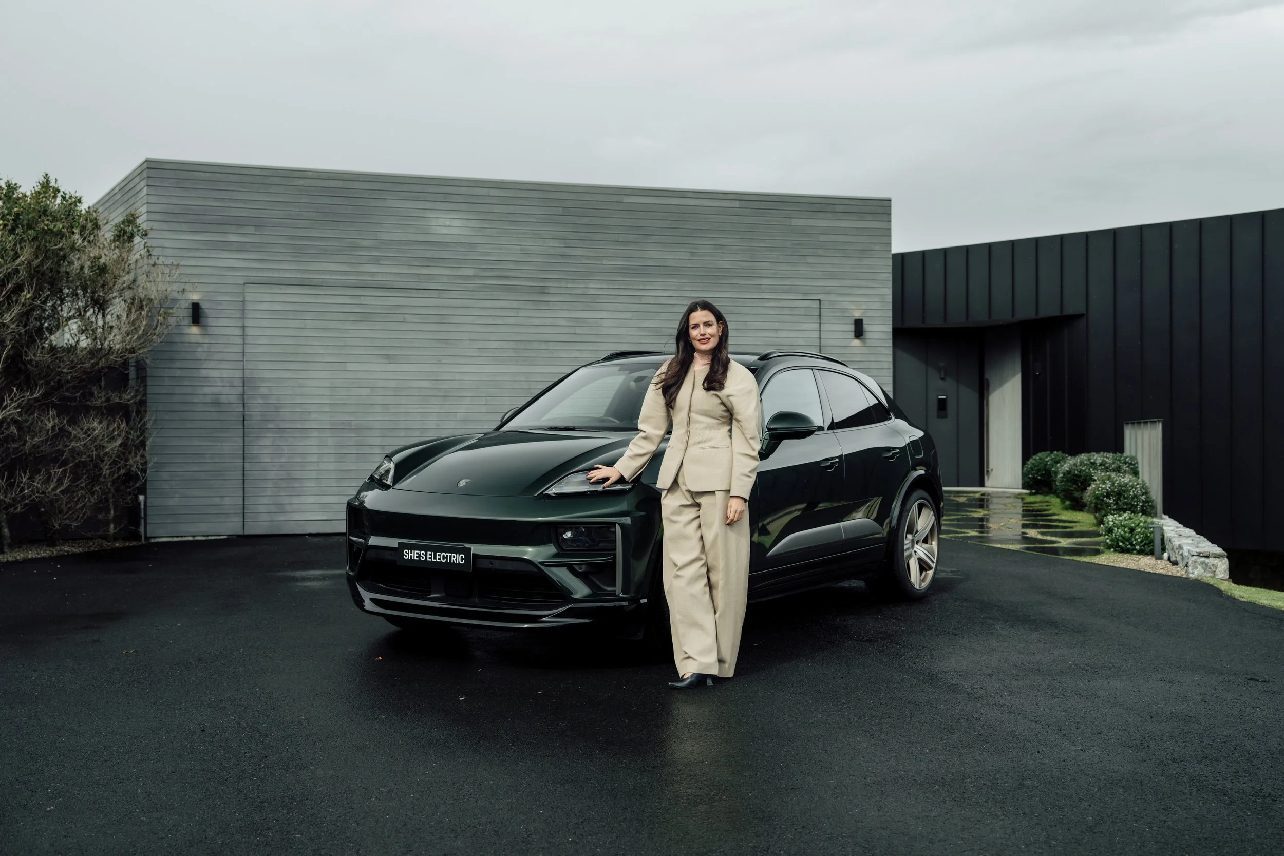 Woman in beige suit standing next to a black electric car in front of modern gray and black building on cloudy day.