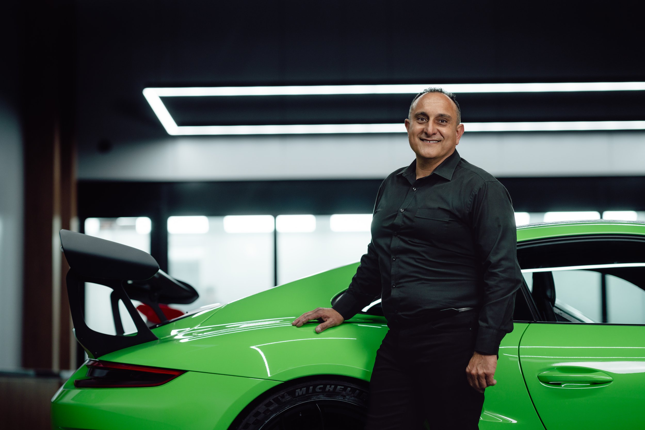 A man in a black shirt standing next to a green sports car inside a modern building with black ceiling and large windows in the background.