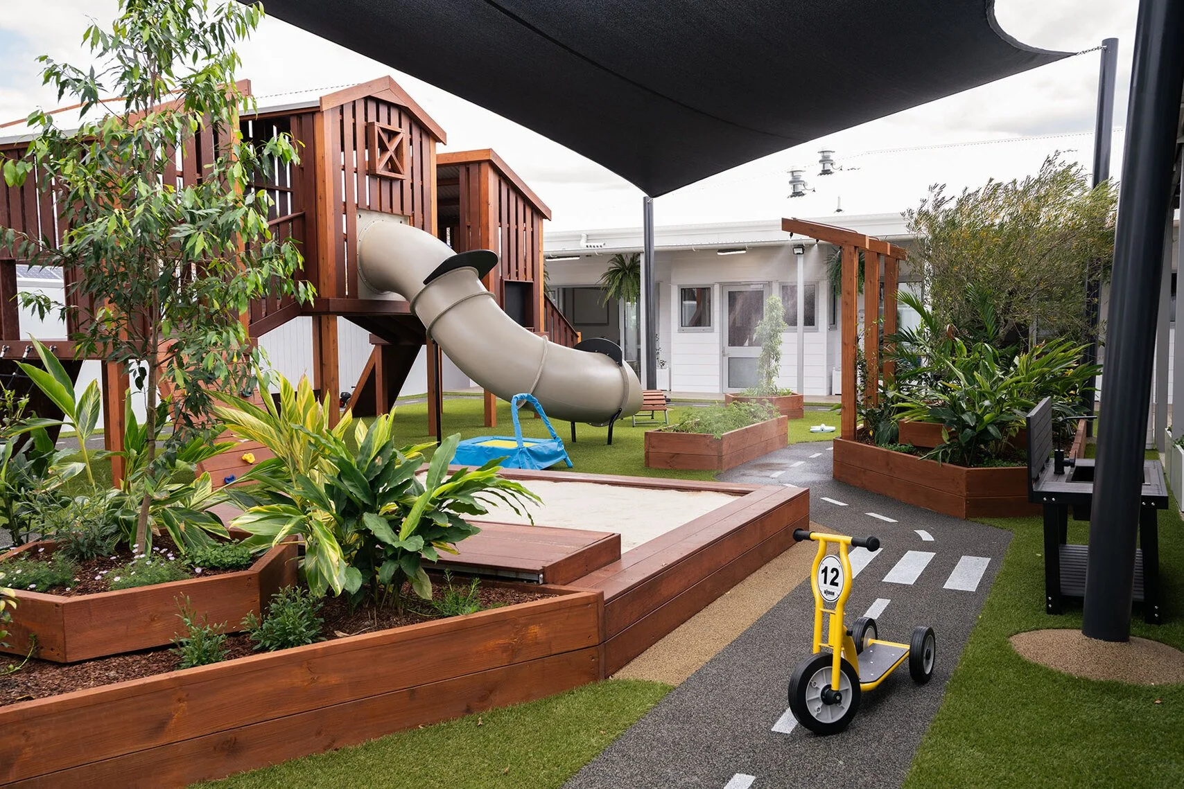 Childrens' outdoor play area with a large wooden play structure, a slide, sandbox, plants, bench, and a small yellow balance bike on a paved path.