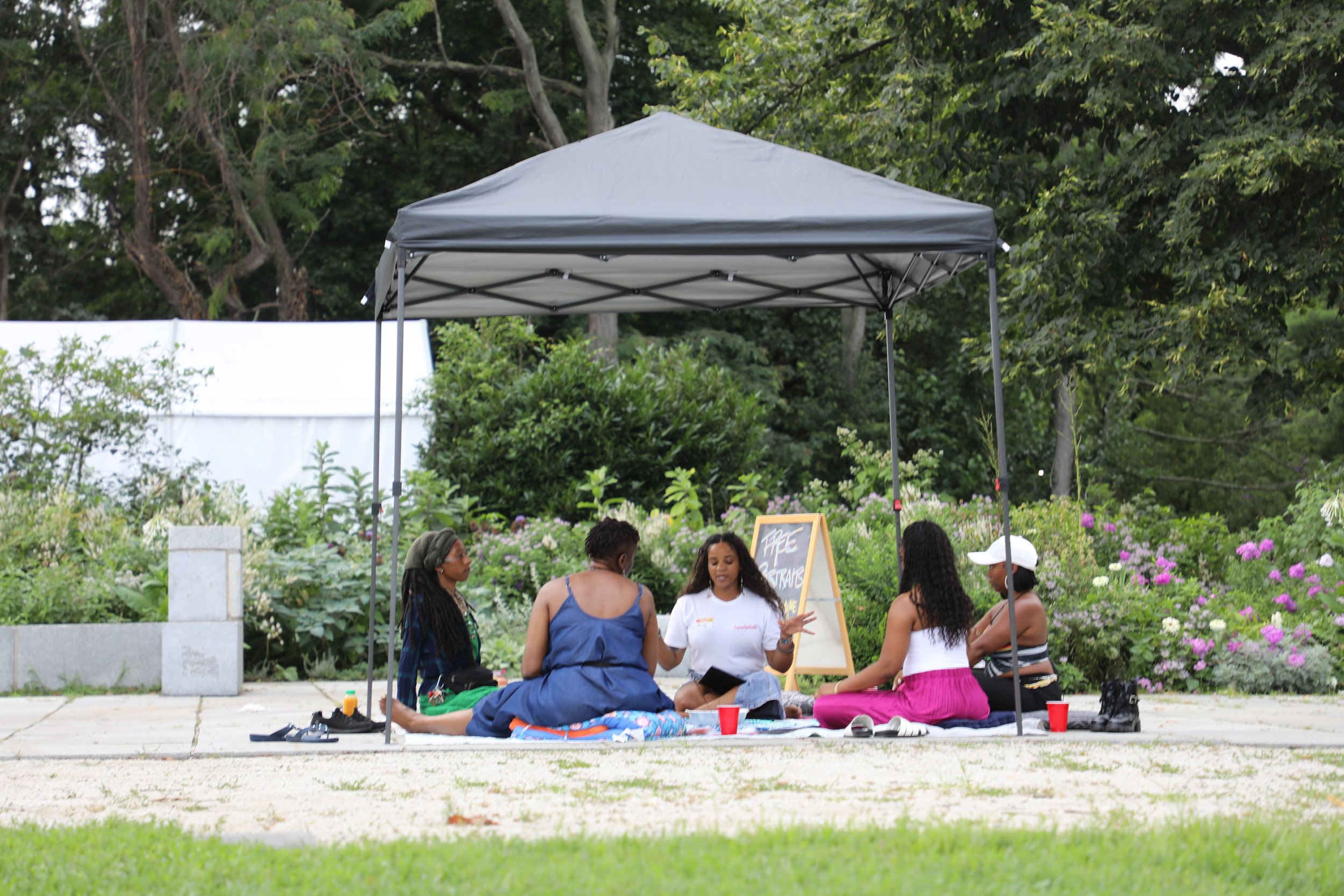 A group of people sitting and chatting on a blanket under a canopy in a garden with trees and flowers.
