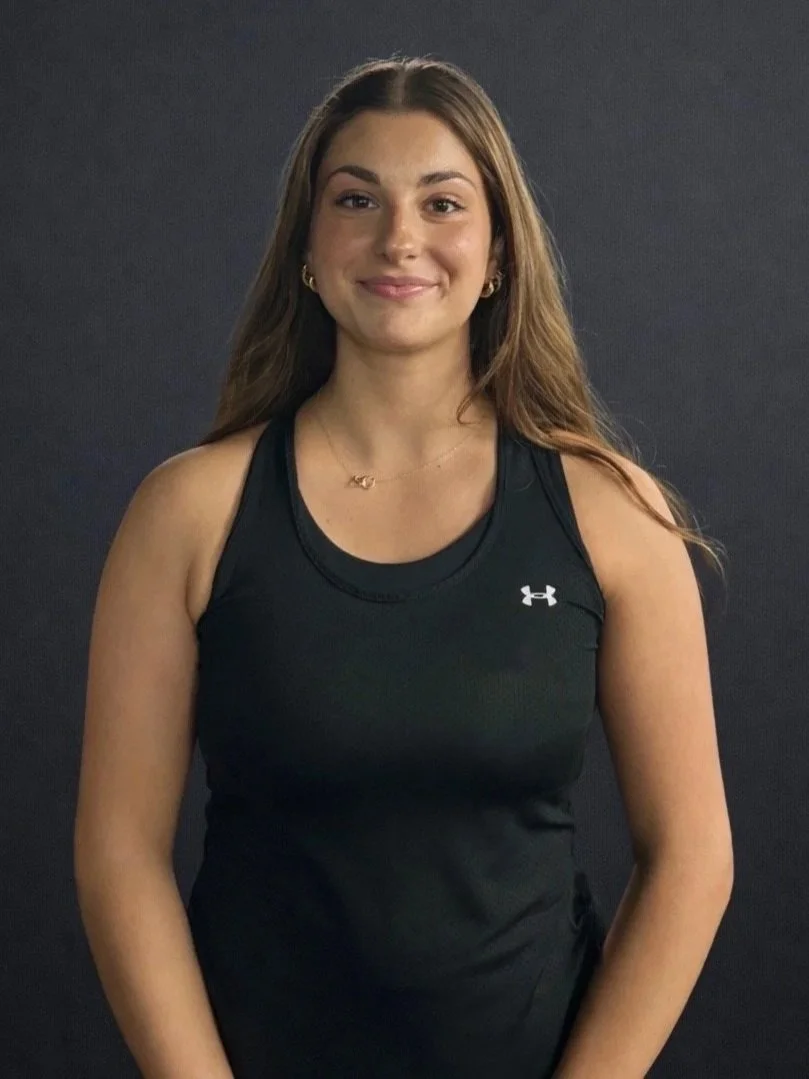 A young woman with long brown hair, wearing a black athletic tank top with Under Armour logo, standing against a dark background, smiling at the camera.