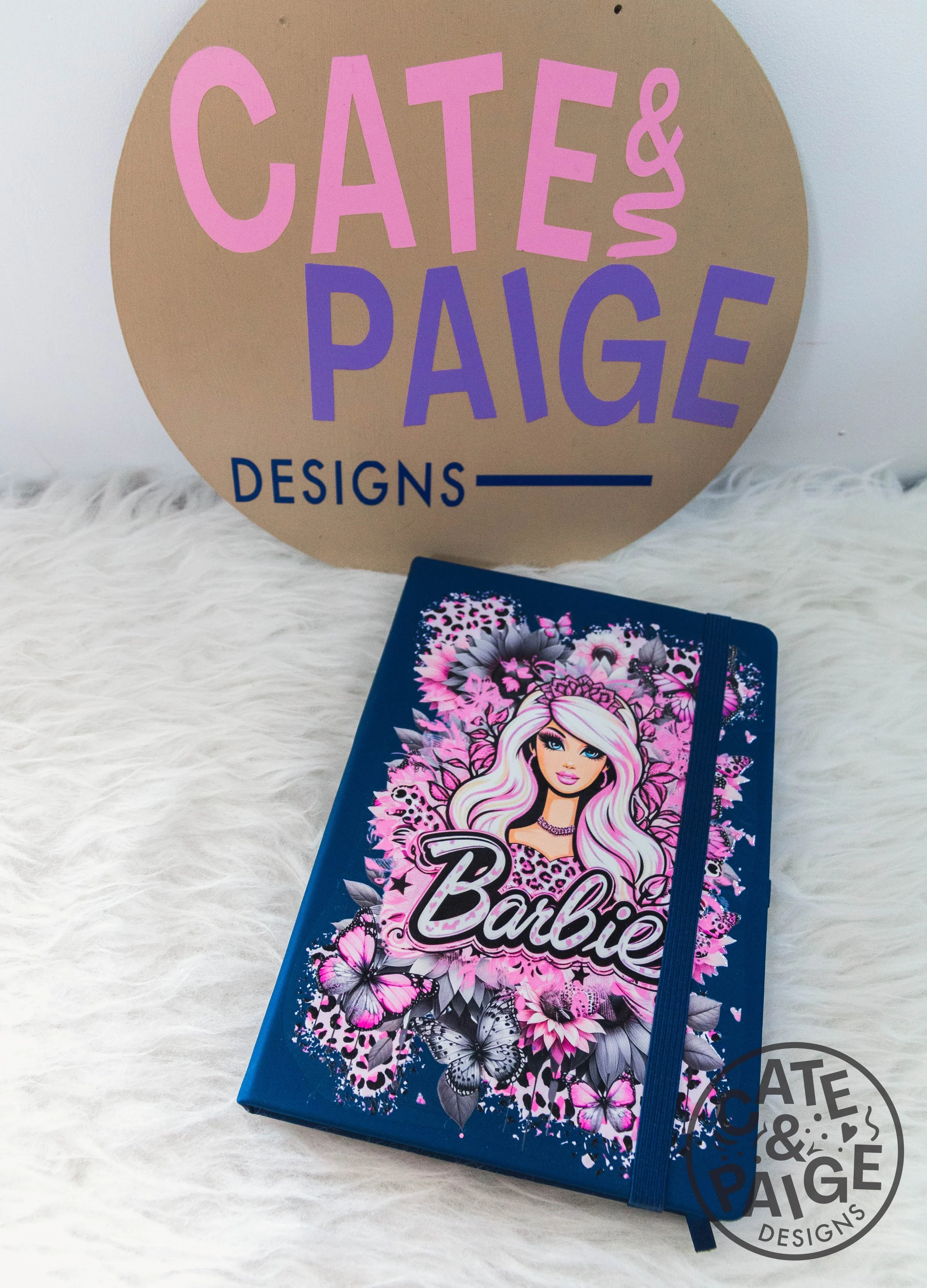 A notebook with a Barbie design on the cover, featuring a blonde Barbie doll with pink, purple, and black floral and butterfly graphics. The notebook has a blue elastic band around it. In the background, there is a round, beige sign with pink and purple text that reads 'CATE & PAIGE DESIGNS.' The items are placed on a white fluffy surface.