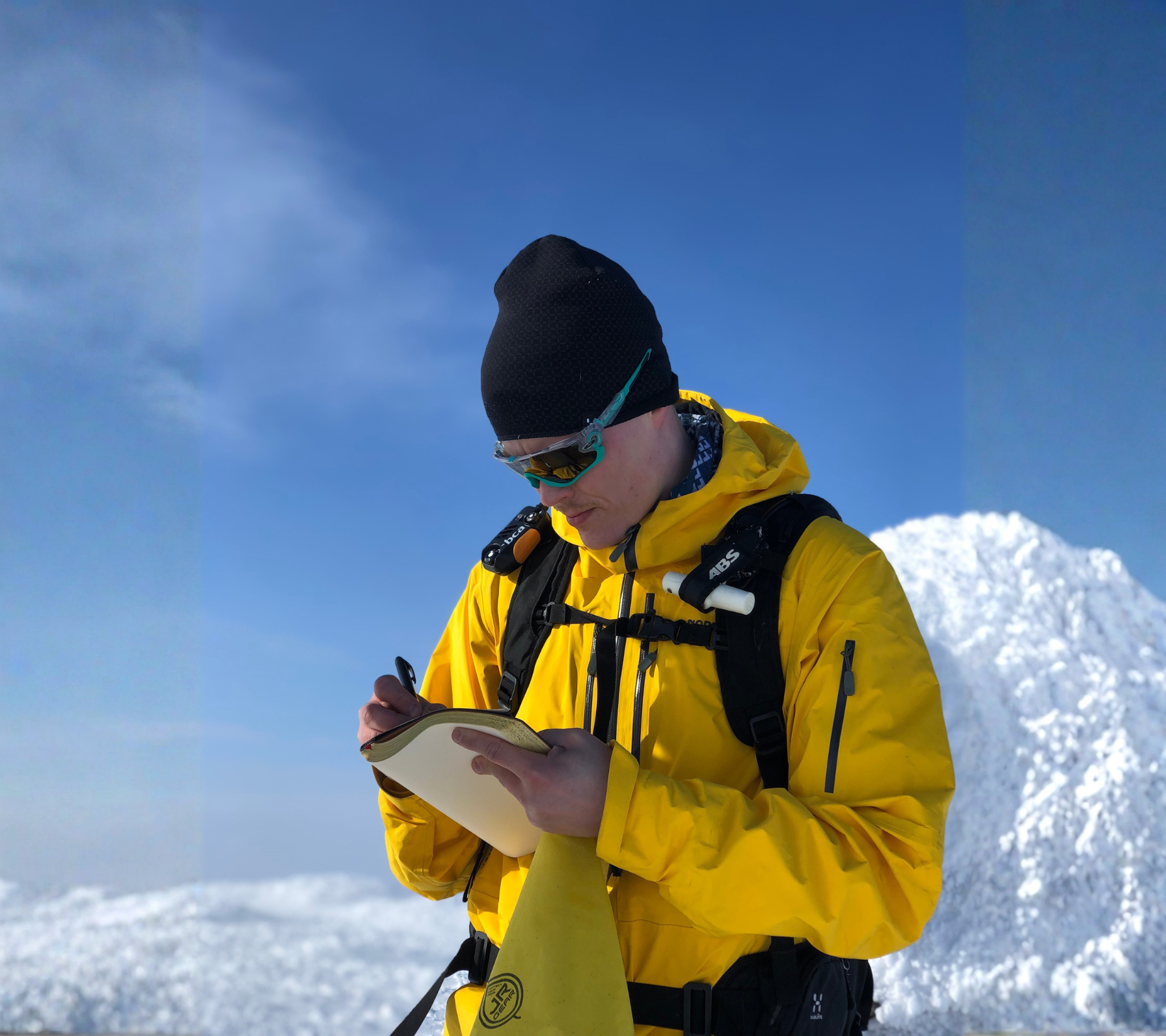 A person in yellow outdoor gear, black beanie, and sunglasses, standing in snowy mountain landscape writing in a notebook.