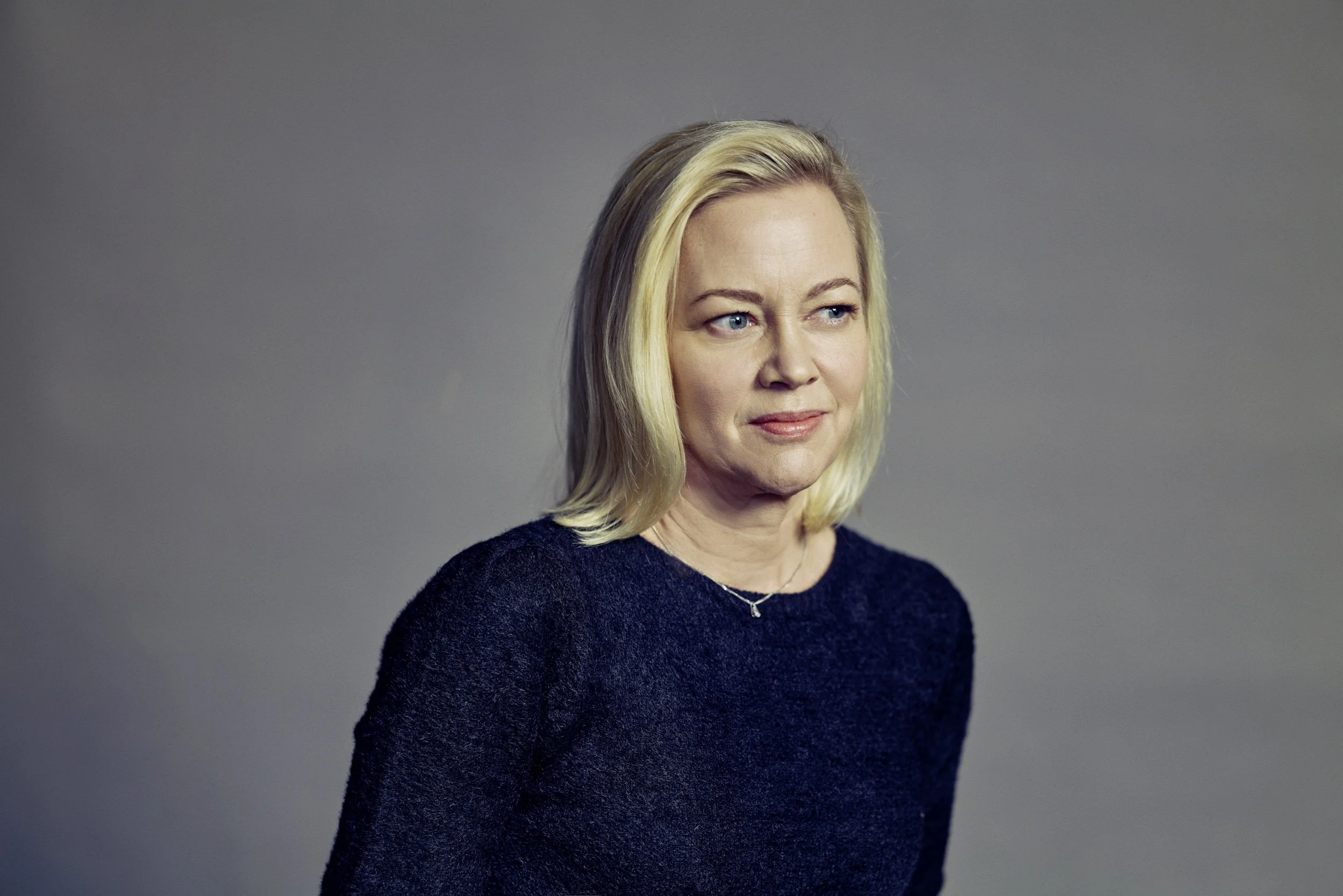 Porträt einer Frau mit blonden Haaren in einem dunklen Oberteil vor grauem Hintergrund.