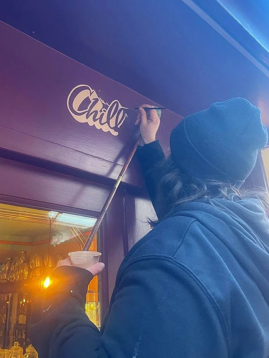 A person wearing a beanie and a hoodie is painting the word 'Chill' on a purple and blue wall with a paintbrush.