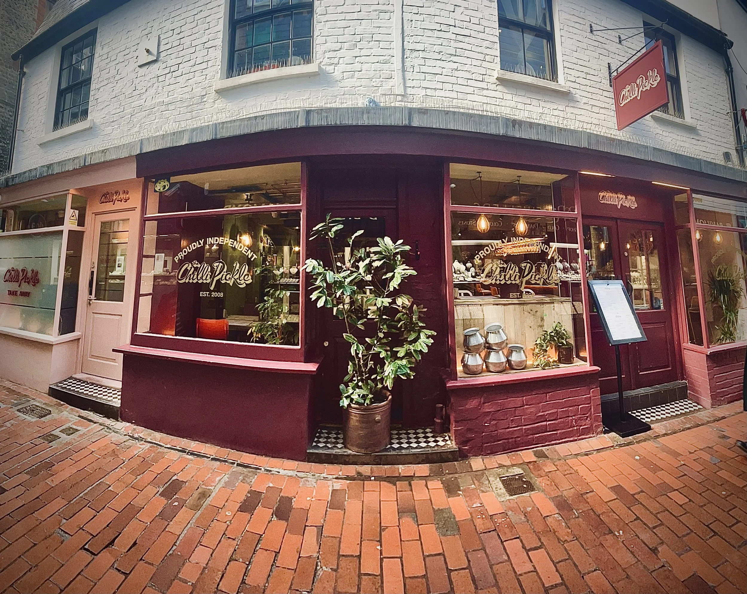 Exterior of a restaurant called Chilli Pickle with a maroon facade, large windows, and a chalkboard menu outside. The sign on the window indicates they are proudly independent, established in 2008. A potted plant is in front of the window, and the si