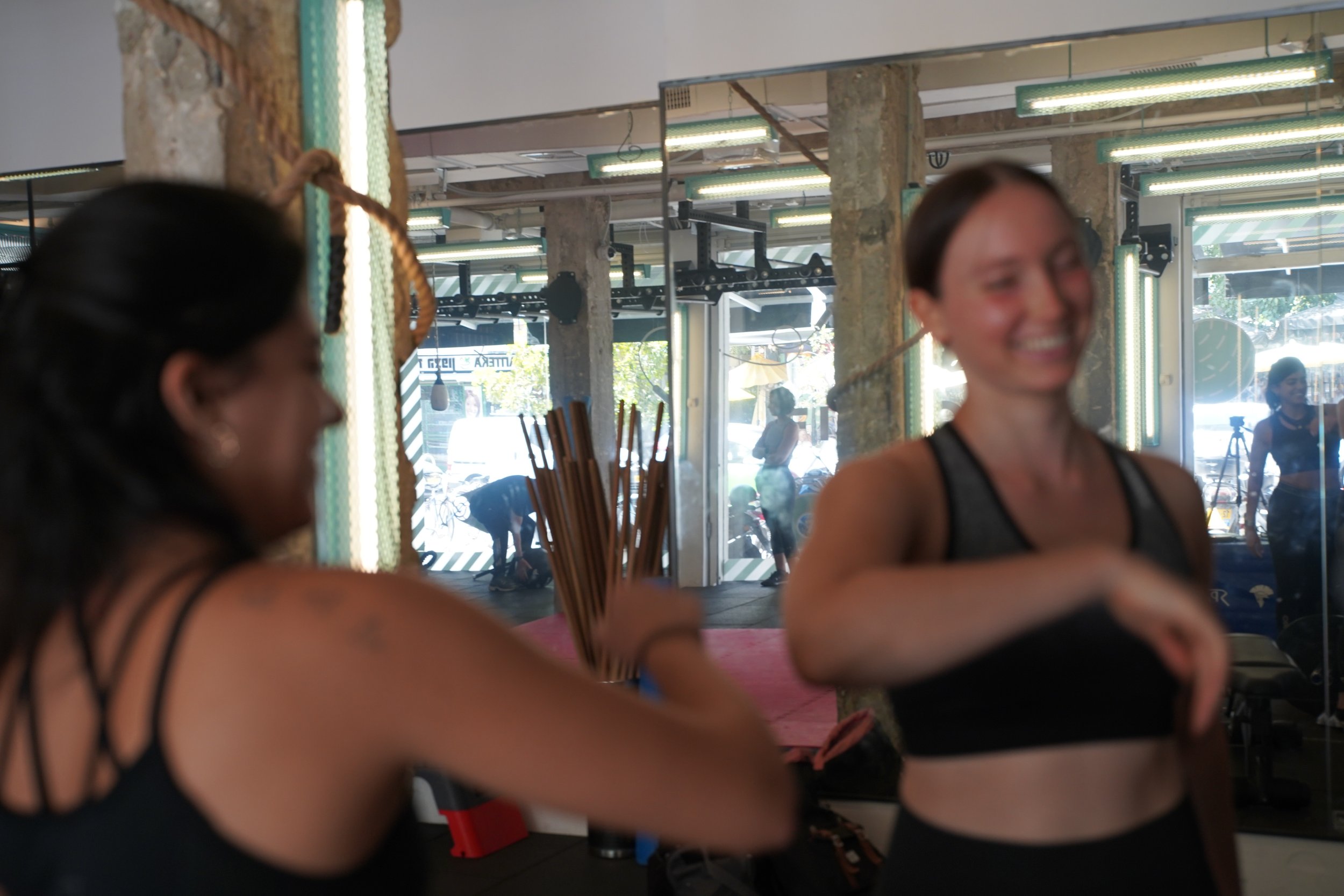 Two women in workout clothes smiling and talking inside a fitness studio with gym equipment and large windows in the background.