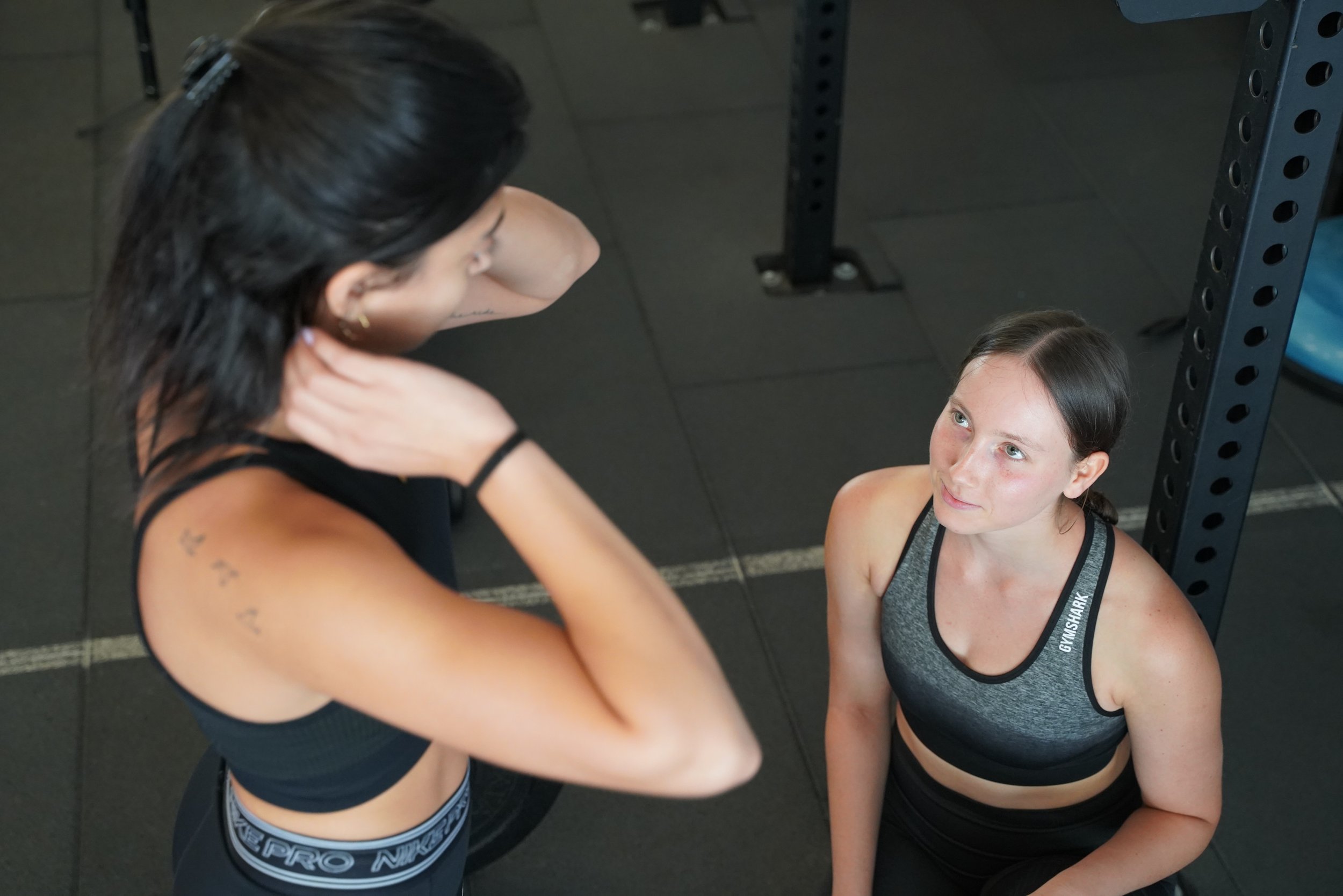 A woman with dark hair in a ponytail is talking to a woman with brown hair tied back at a gym. They are both dressed in athletic wear, and the woman with brown hair is sitting on the gym floor looking up at the standing woman. Exercise equipment is visible in the background.