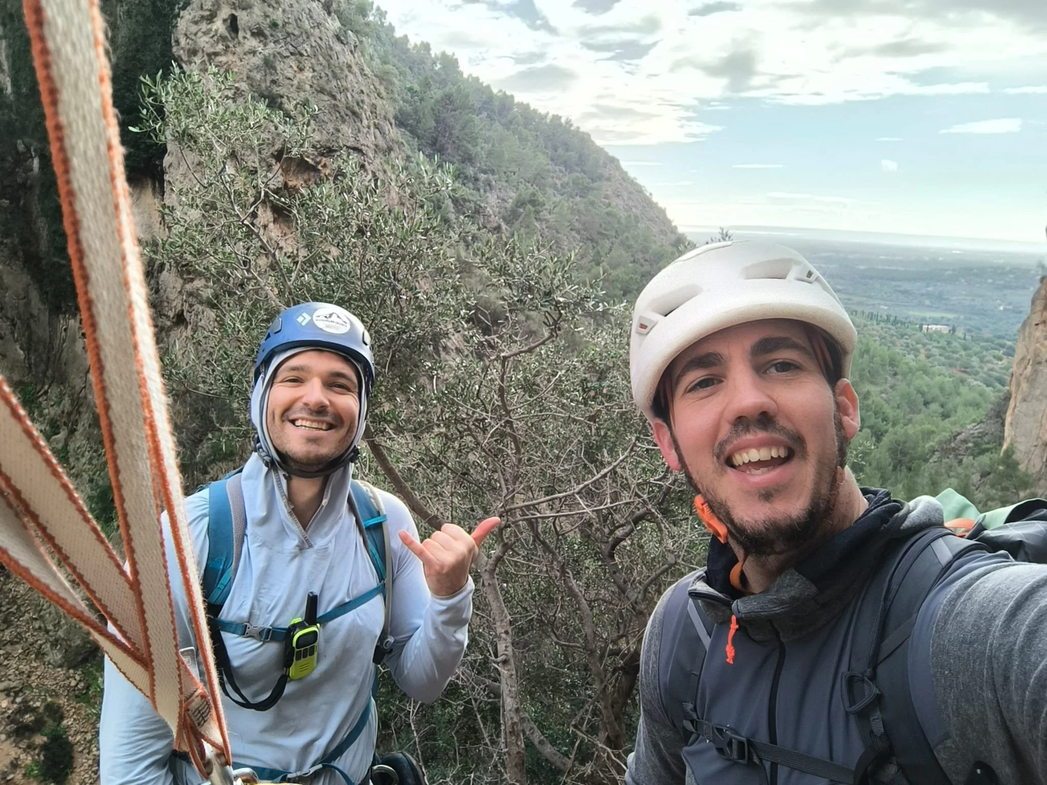 Two men wearing helmets and backpacks taking a selfie outdoors on a mountain trail with trees and a valley in the background.