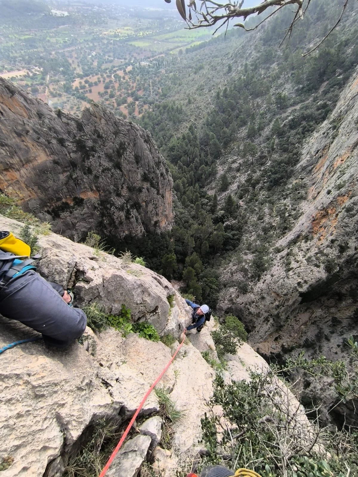 Climbers ascending a steep rock face on a mountain, with a valley and forest in the background.