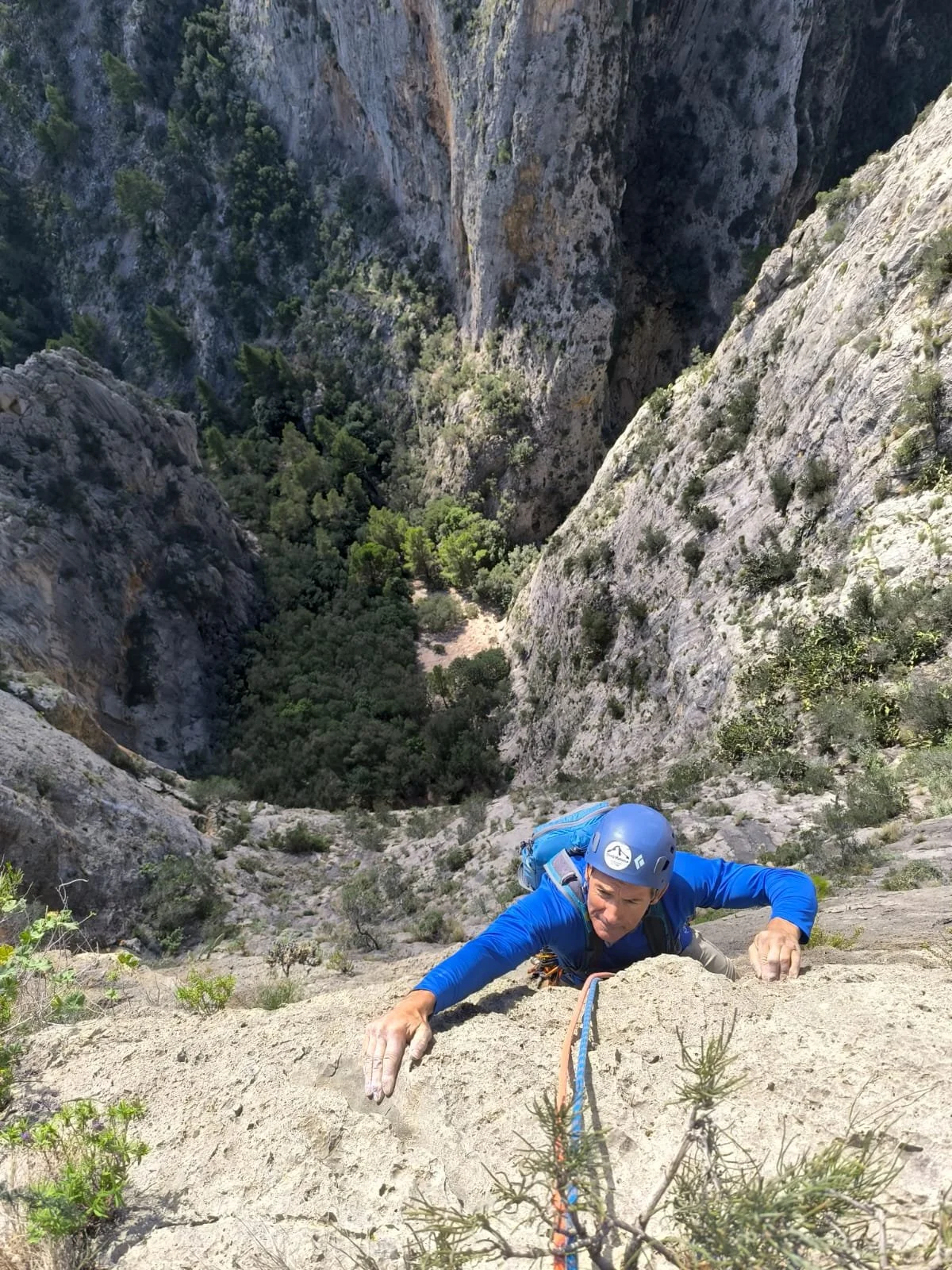 A person rock climbing on a steep cliff face, wearing a blue jacket, helmet, and harness, with a view of a deep valley below.