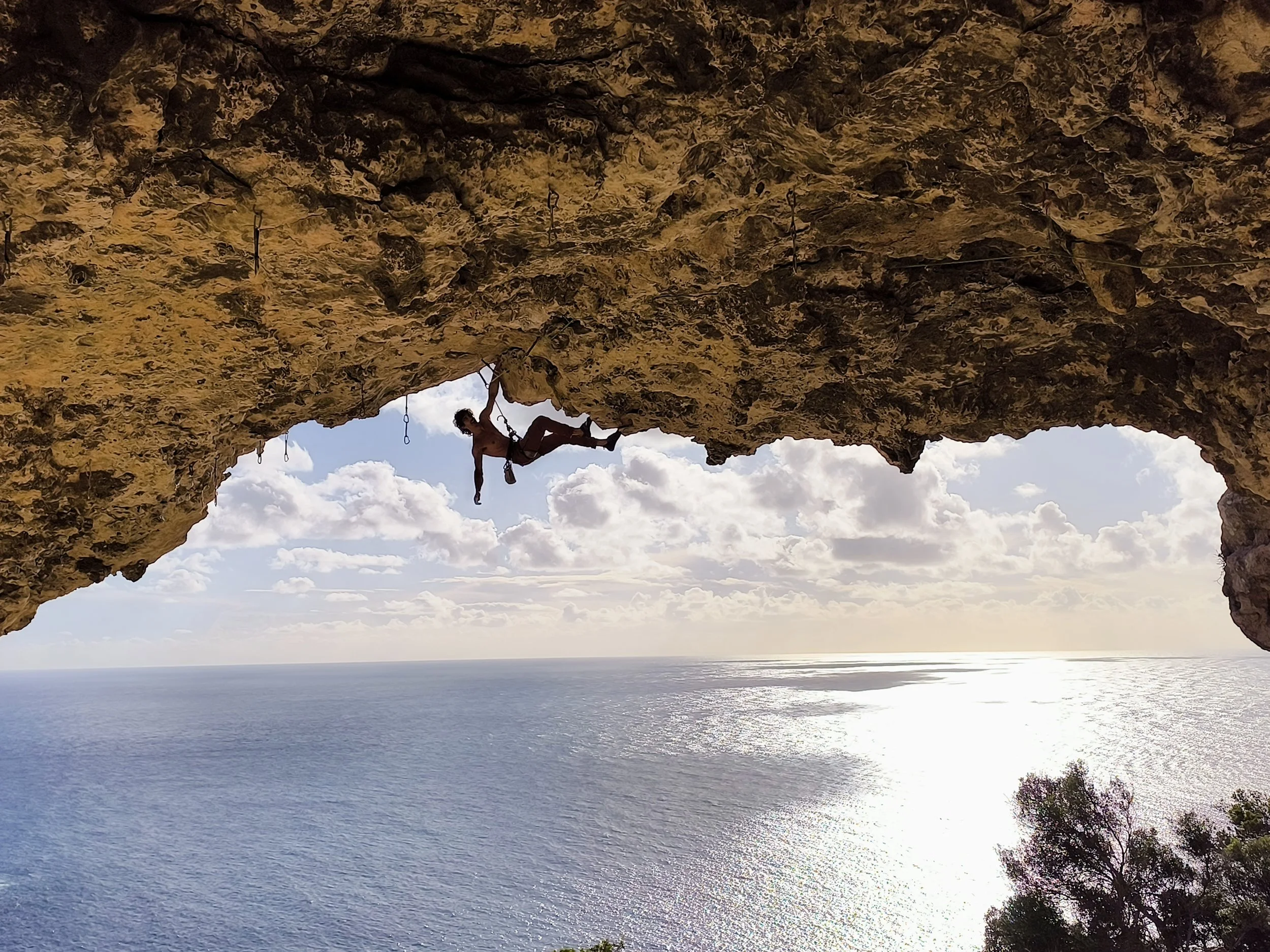 A person is rock climbing on a large overhanging cliff above the ocean during sunset, with clouds and trees in the background.
