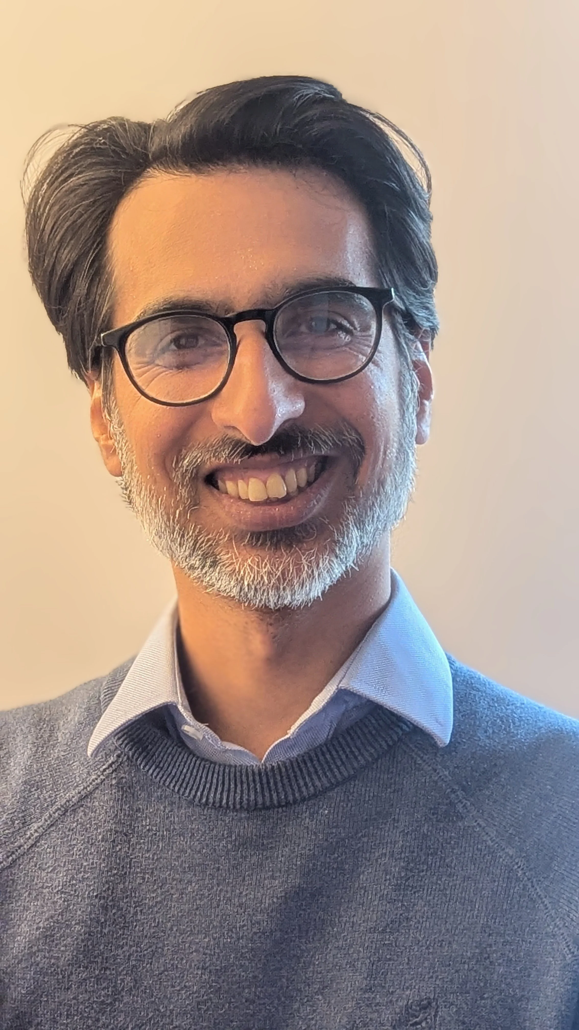 A middle-aged man with glasses, a beard, and gray hair, smiling and wearing a light blue collared shirt and a gray sweater, standing against a plain, light-colored background.