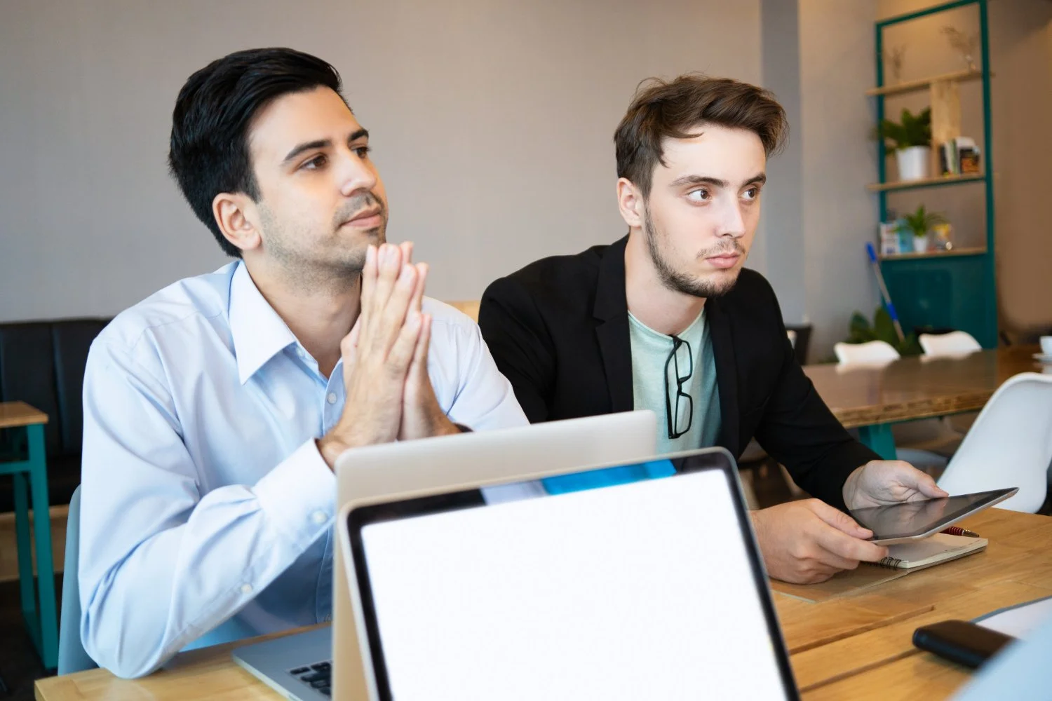 Two men sitting at a desk during a meeting, one with hands clasped in prayer and eyes closed, the other holding a tablet, in a modern office or café setting.