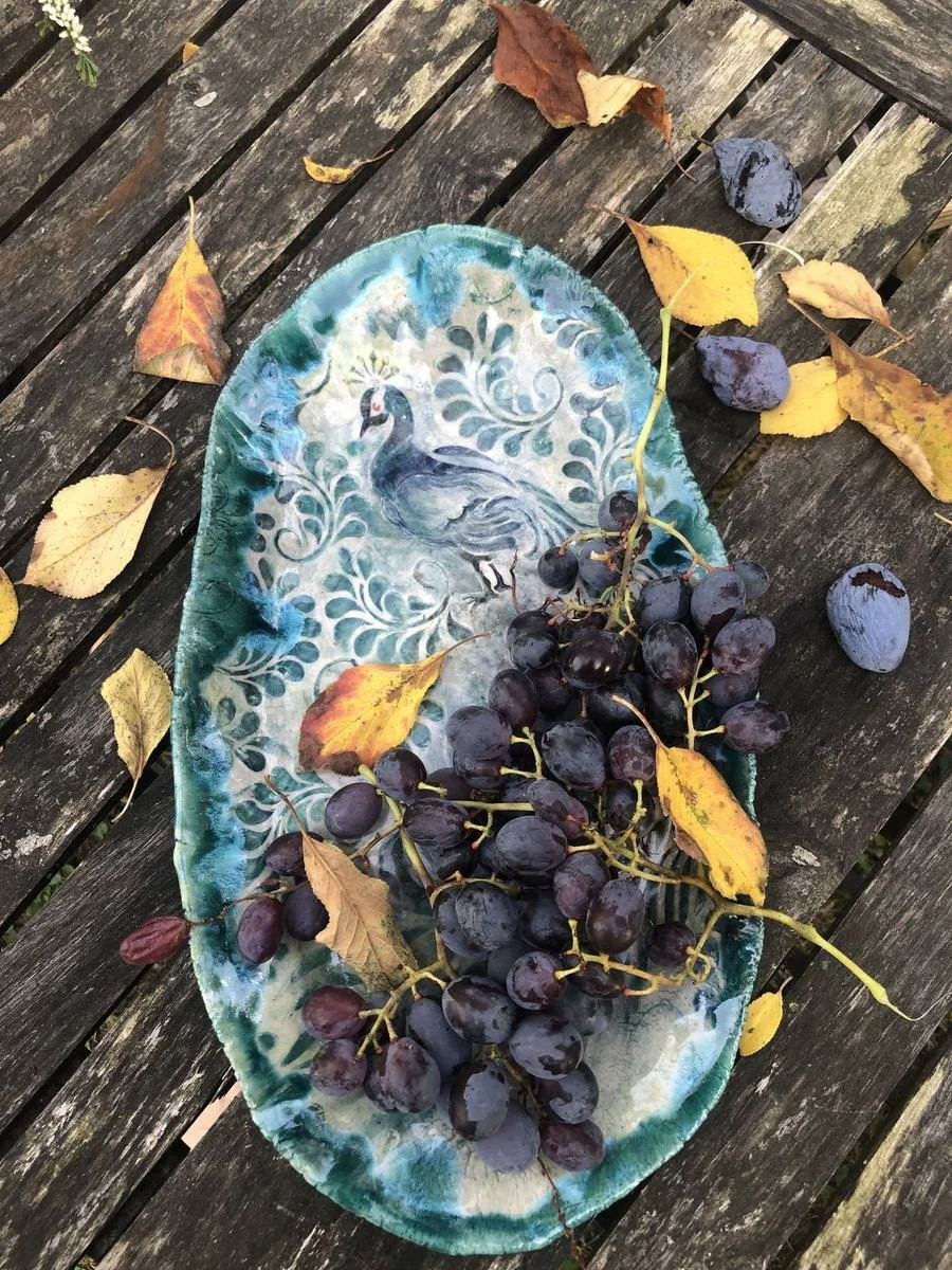Keramikteller mit blauem Vogelmotiv, darauf dunkelblaue Weintrauben und Herbstblätter, auf Holztisch.