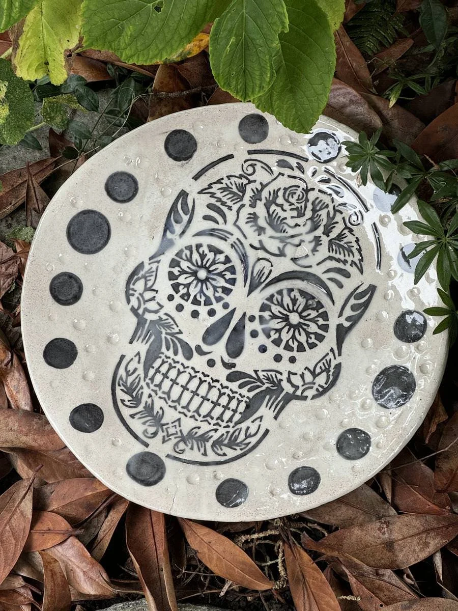 Keramikteller mit schwarzem Totenkopfdesign und Punkten, umgeben von grünen Blättern und braunen Blättern am Boden.