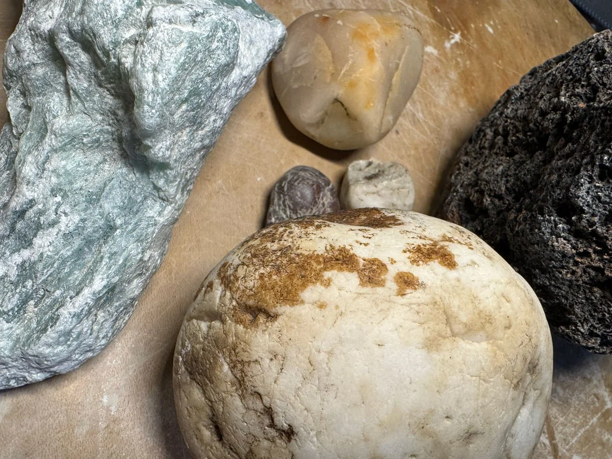 Verschiedene Steine auf einer Oberfläche, darunter ein grünlicher, ein hellbrauner, ein schwarzer poröser und weitere kleinere Steine.