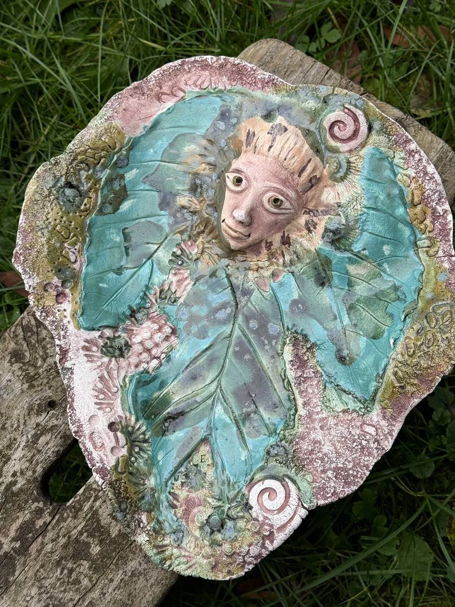 Keramikplatte mit floralen und menschlichen Motiven, bunten Verzierungen und reliefartigen Oberflächen, auf Holzuntergrund im Freien platziert.