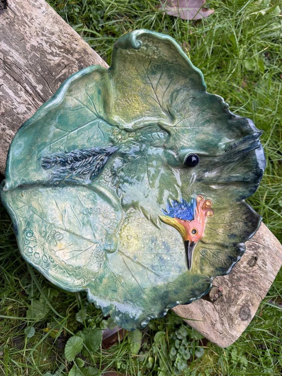 Keramikteller in Form eines grünen Blattes mit einem geschnitzten Vogelkopf in der Mitte, auf einem Holzstück liegend.