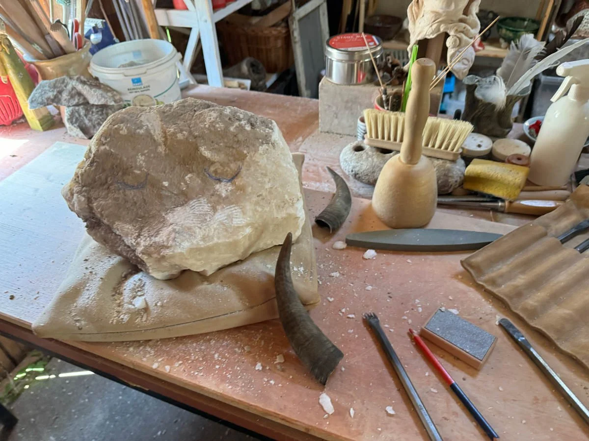 Ein Bild von einer Werkbank mit einem großen Stein, umgeben von verschiedenen Bildhauerwerkzeugen wie Meißeln, Feilen und einem Gummihammer. Es gibt auch zwei Hörner und andere Gegenstände im Hintergrund.