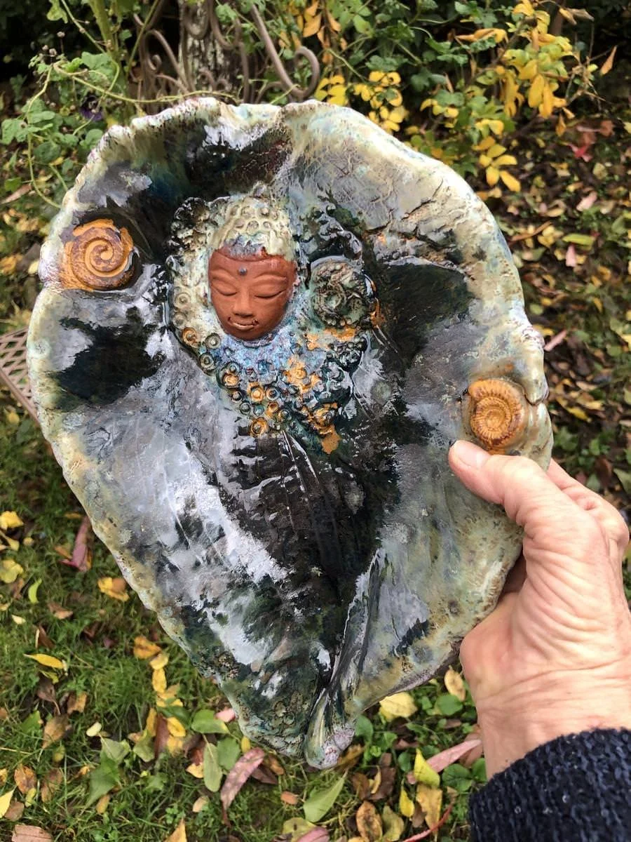 Ein kunstvoll gestaltetes Keramikobjekt in Form eines Blattes mit einem eingearbeiteten Gesicht in der Mitte, das eine meditative Pose zeigt, gehalten von einer Hand. Es ist von aufwendigen Mustern und Spiralformen umgeben, alles in natürlichen, erdi