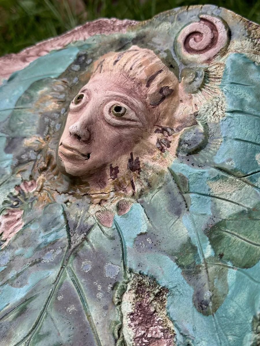 Keramikskulptur mit einem Gesicht, das von Blattmustern umgeben ist. Die Farben sind hauptsächlich Türkis und Beige mit einigen rostigen Akzenten.