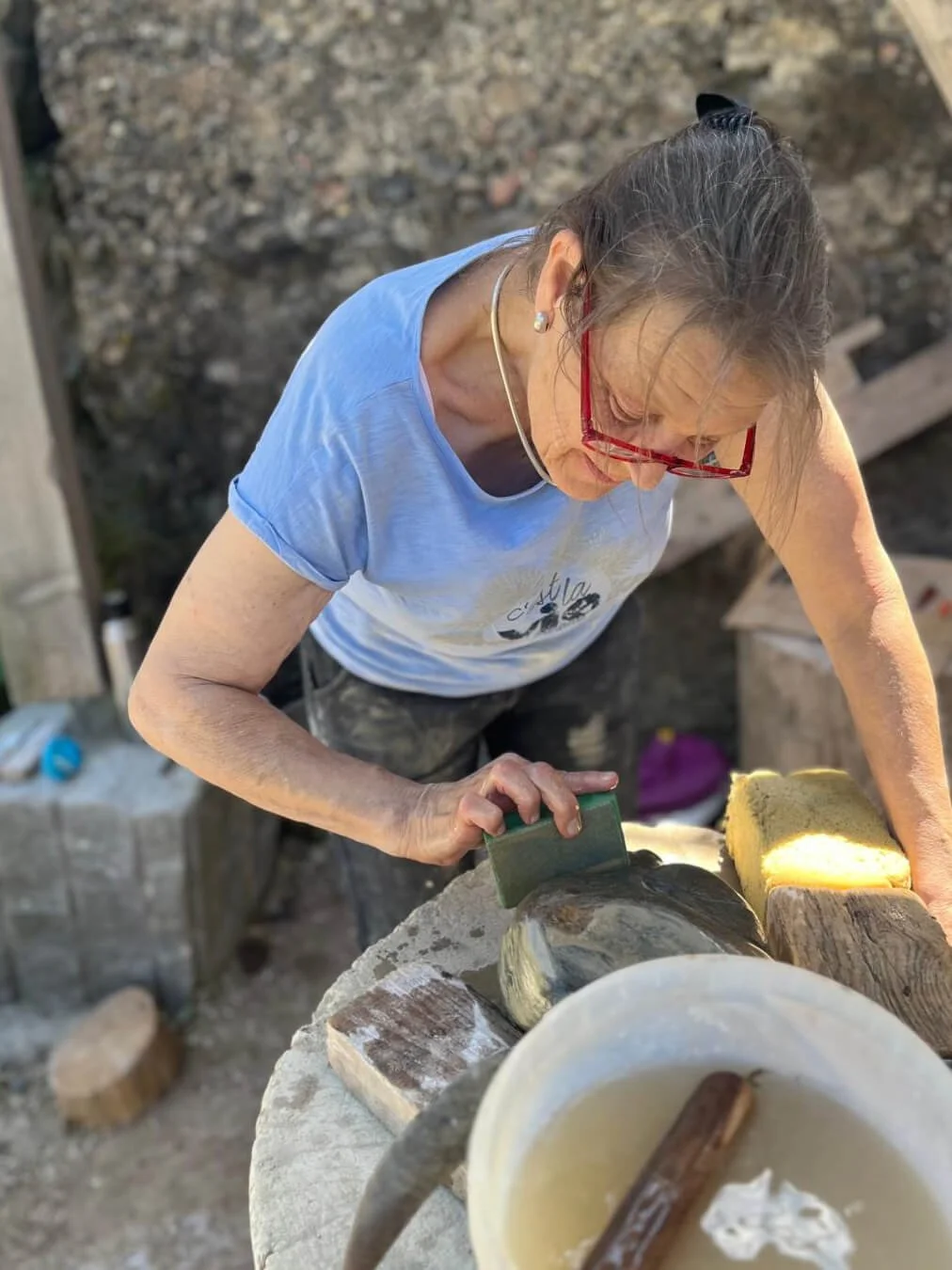 Eine Frau bearbeitet mit einem Schleifwerkzeug eine Skulptur aus Stein oder Ton auf einem Tisch im Freien. Neben ihr steht ein Eimer mit Wasser und ein Schwamm.