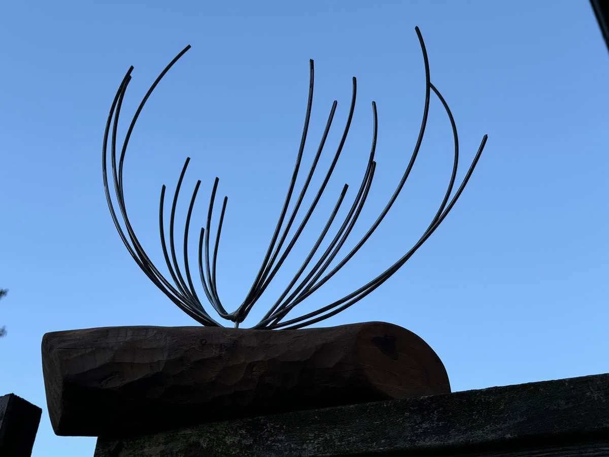 Metallskulptur mit geschwungenen Drähten auf einem Holzblock vor einem blauen Himmel.