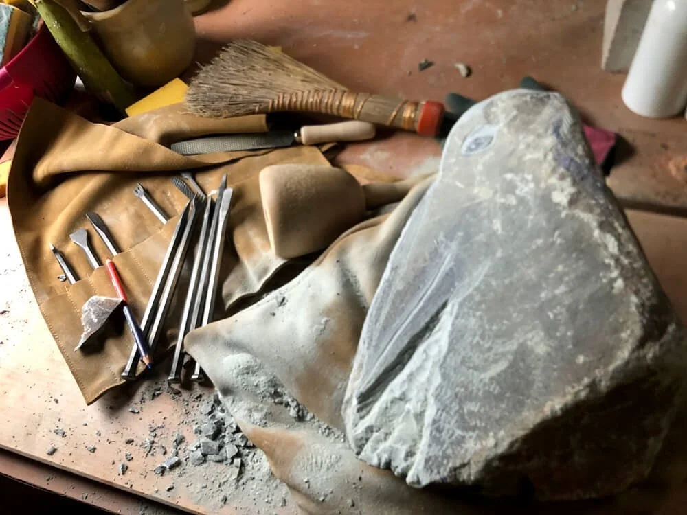 Werkzeuge für Steinbearbeitung auf einem Tisch, darunter Feilen, Meißel und Bürste, neben einem grob bearbeiteten Stein.