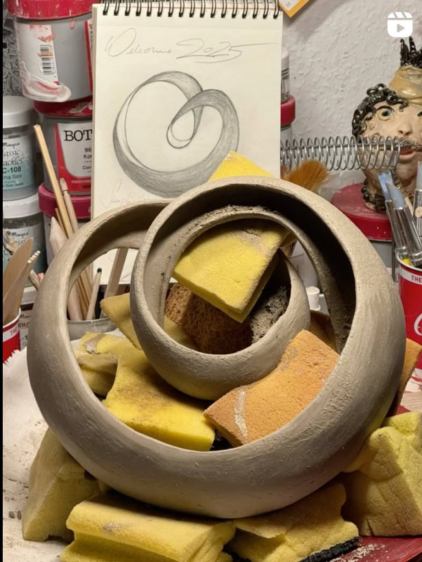 Töpferton Skulptur mit gelben Schwämmen und Skizze im Hintergrund, umgeben von Werkstattmaterialien.