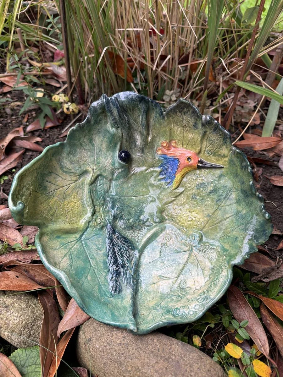 Dekorativer Keramikteller in Form eines grünen Blattes mit einem bunten Vogel, umgeben von Pflanzen und Blättern im Freien.