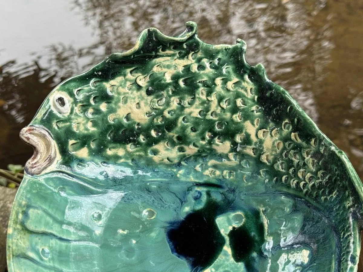 Keramikteller in Form eines grünen Fisches gehalten, auf einem Hintergrund aus Wasser. Der Teller hat eine strukturierte Oberfläche mit Details, die Schuppen und Flossen eines Fisches darstellen.