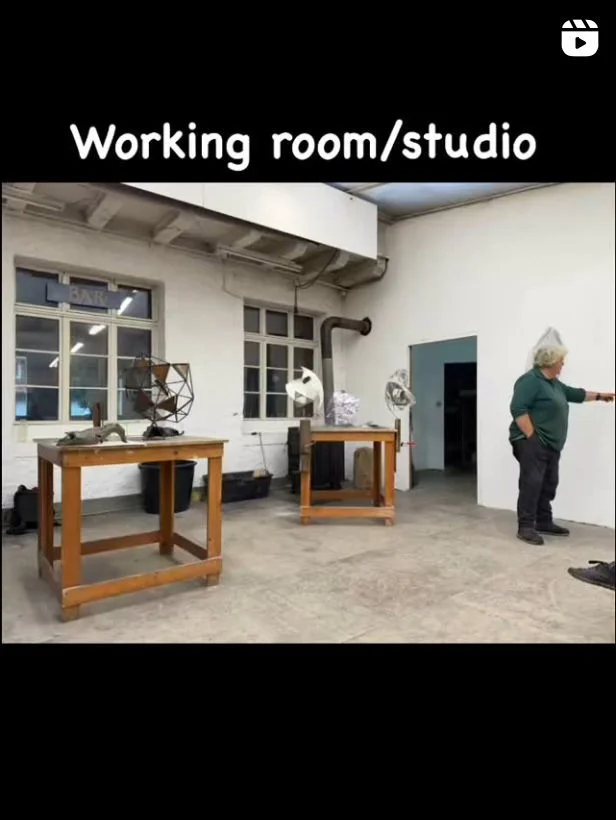 Werkraum mit weißen Wänden und großen Fenstern, zwei Holztische mit Metallobjekten darauf. Eine Person zeigt an die Wand.