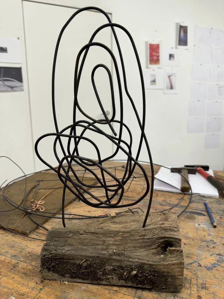Eine abstrakte Skulptur aus dünnen schwarzen Drahtschlingen, die auf einem groben Holzblock montiert ist, steht auf einem Tisch in einem Atelier oder Arbeitsraum. Im Hintergrund sind Werkzeuge und Notizen an der Wand zu sehen.