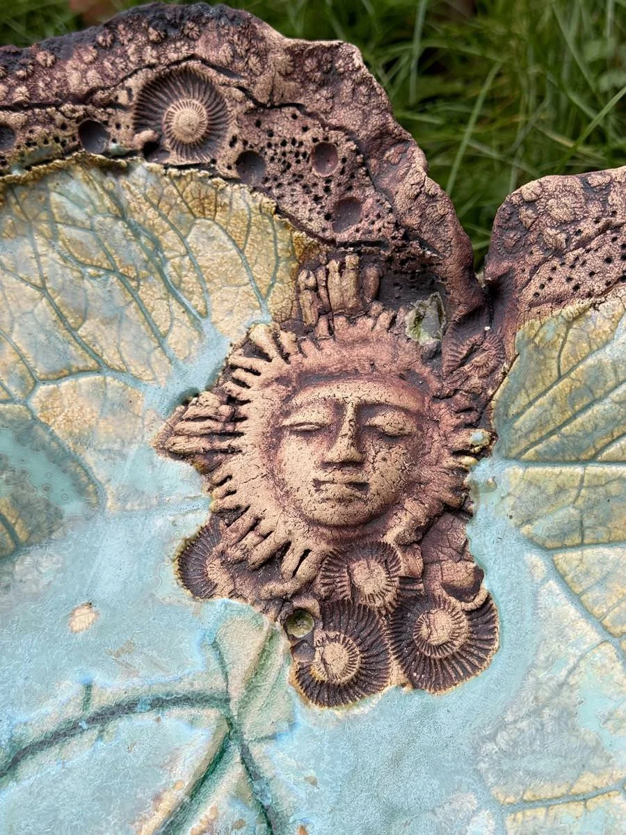 Keramikskulptur mit einem Sonnengesicht und Muschelverzierungen, farbig in Braun und Grün. Das Motiv ist auf einer strukturierten Oberfläche angebracht, umgeben von pflanzenartigen Mustern.