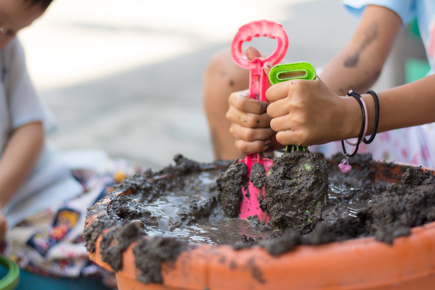 child making mud pie in large terracota pot. A spade in both hands (one green one pink). Lots of mud