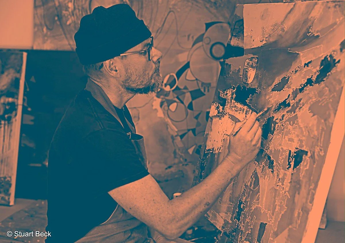 The Artist Stuart Beck artist is painting an abstract canvas with various shades, while working in a studio with colourful artwork in the background.