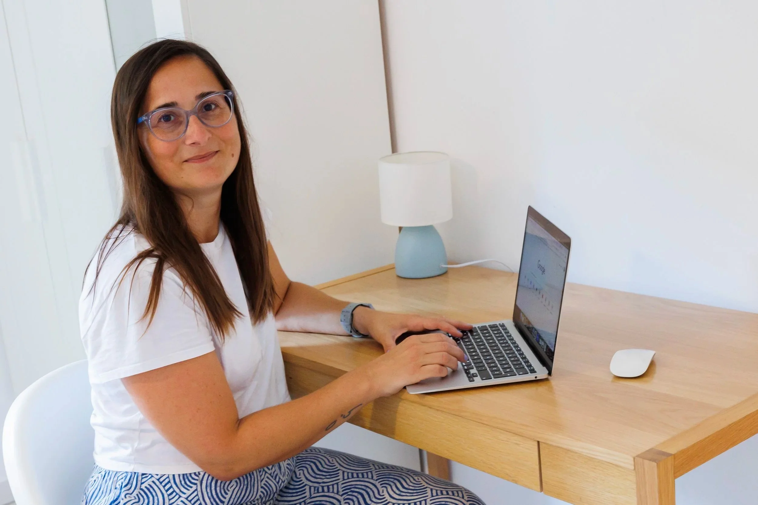 Woman with glasses sitting at a wooden desk using a laptop, with a small white lamp on the desk.