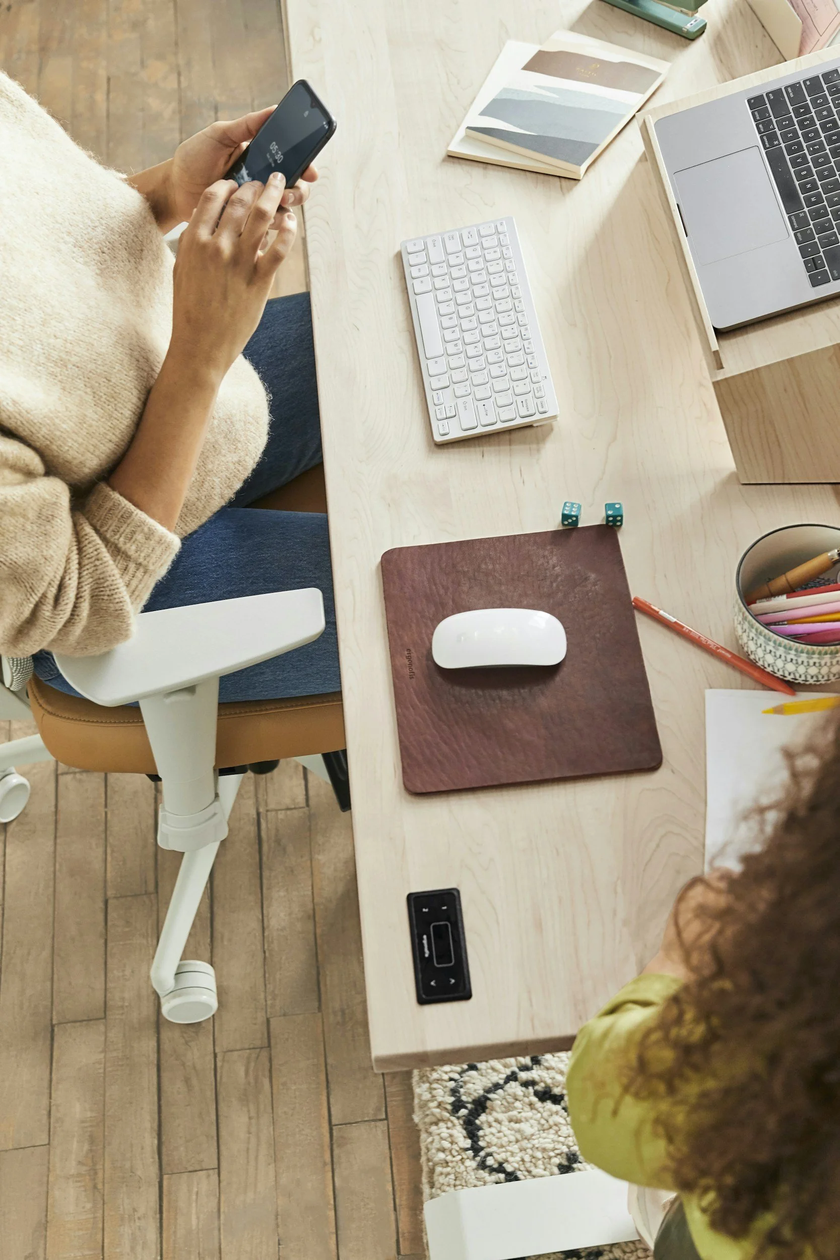 Overhead view of a workspace with two people. On the desk: a laptop, a wireless keyboard, dual small blue dice, a white mouse on a brown mouse pad, a cup with pens and pencils, some papers, and a book. One person is holding a smartphone. The workspace has wooden flooring and a beige office chair.