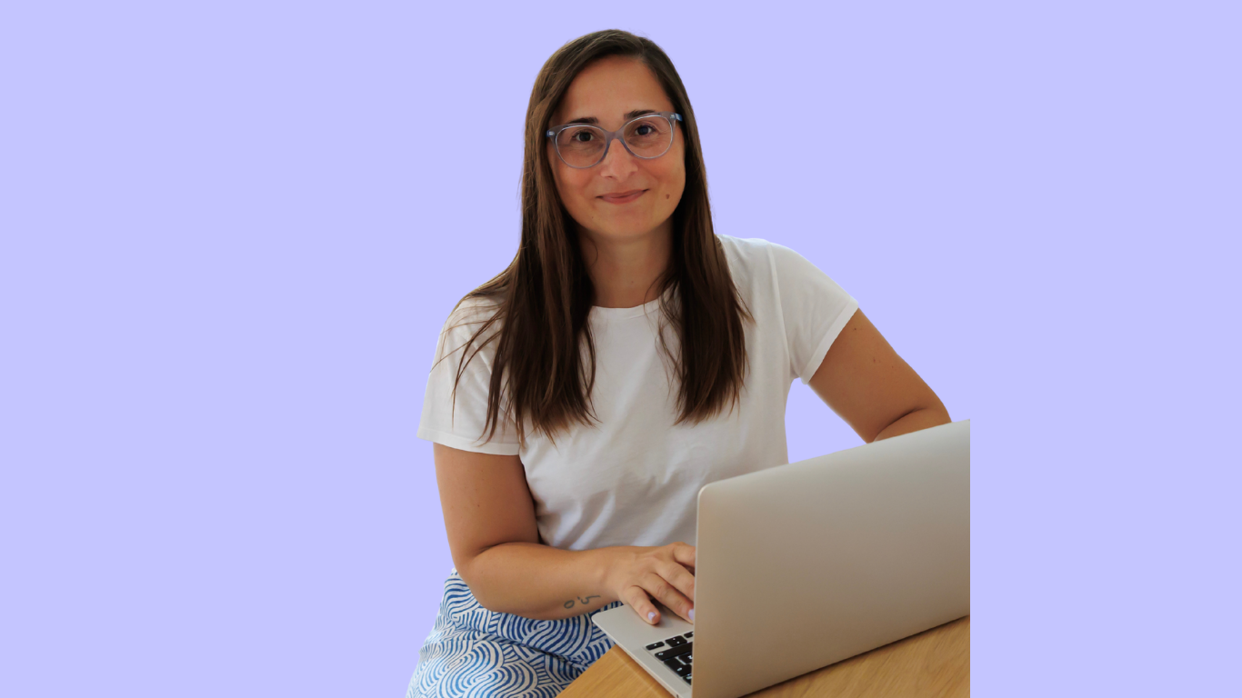 Woman with glasses working on a silver laptop at a wooden table against a light purple background.