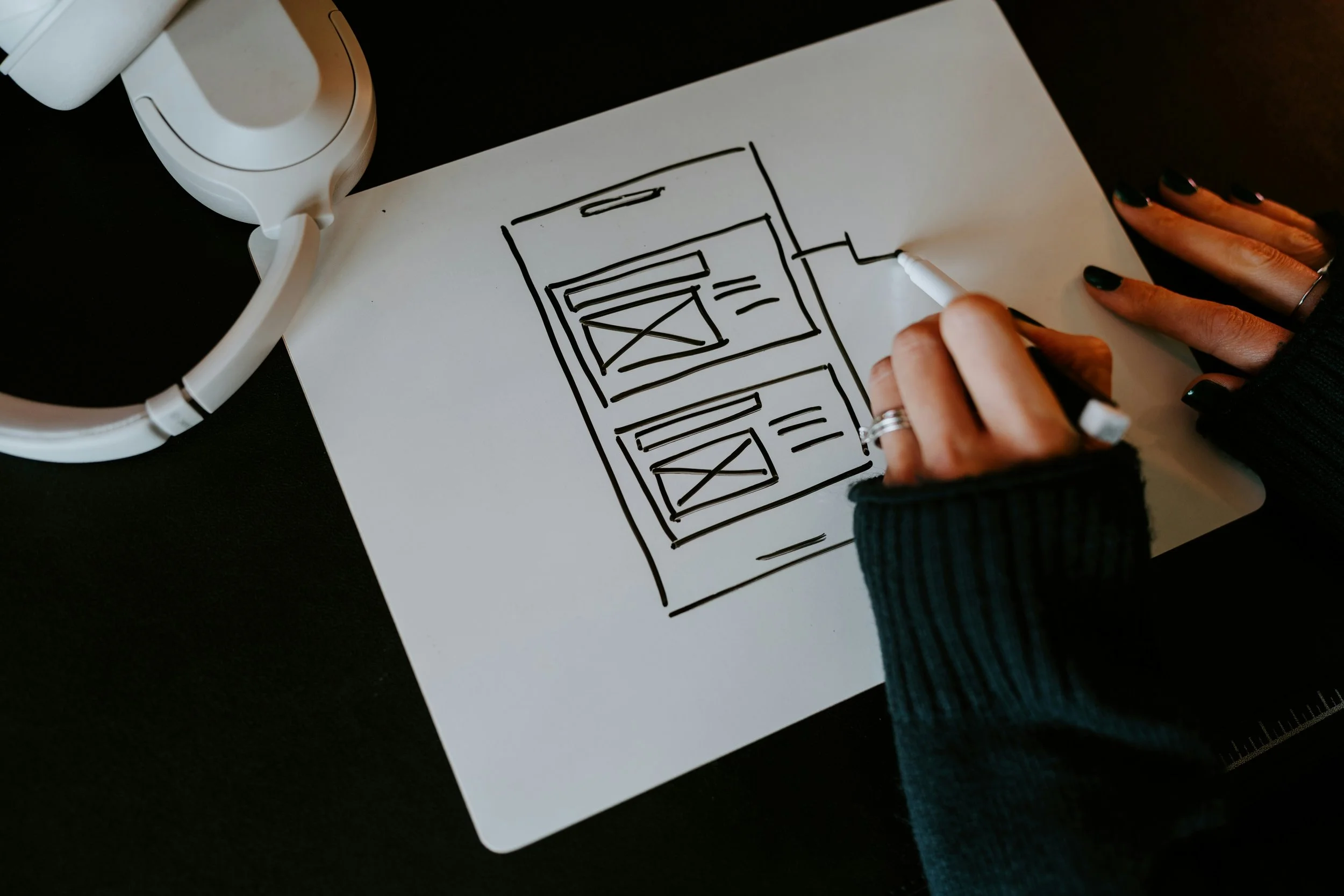 A person sketching a wireframe layout of a mobile app on a white sheet of paper with a black marker, with headphones nearby on a black surface.