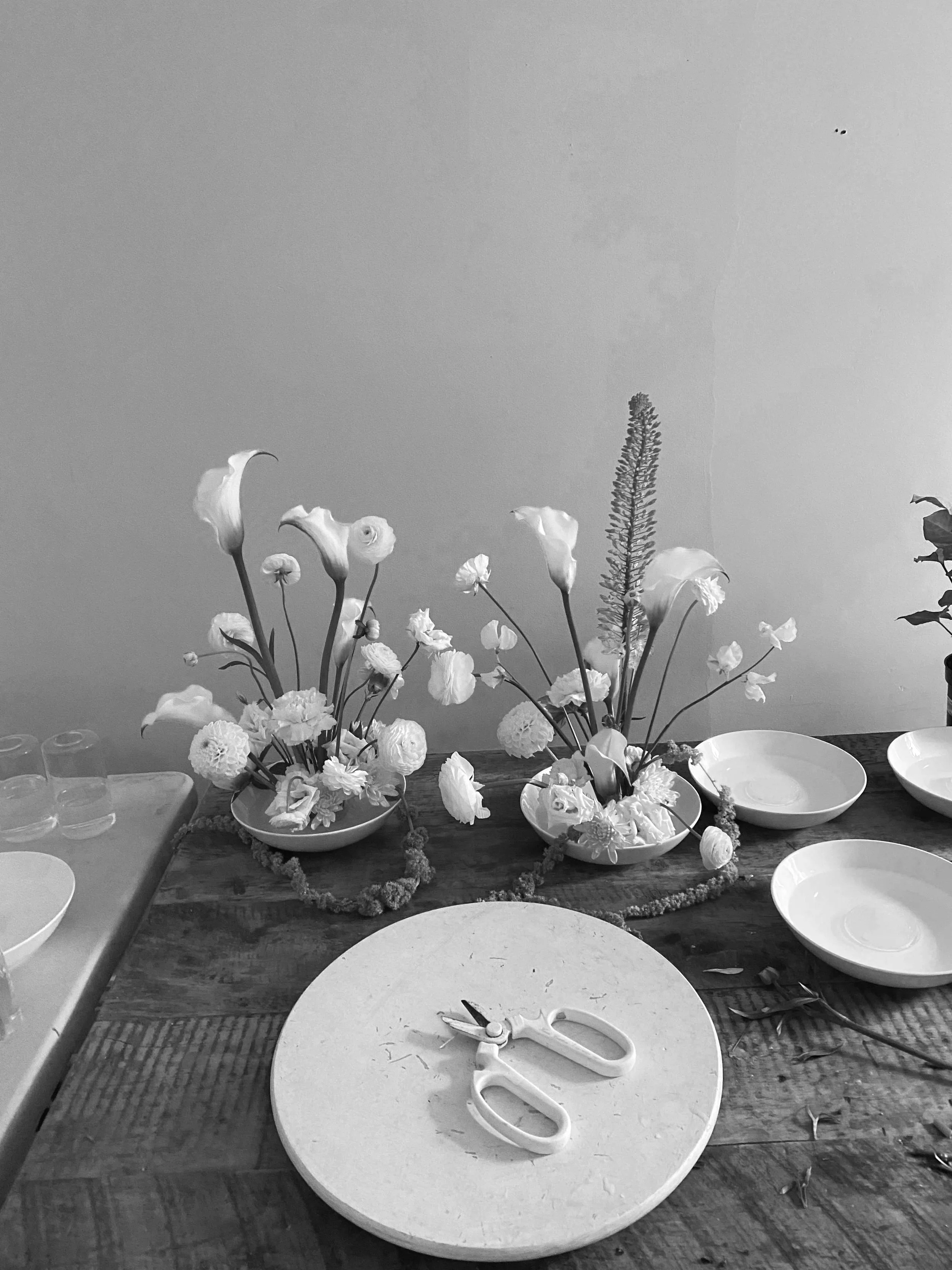 A black and white photo of a table with floral arrangements in bowls, a pair of scissors, and empty bowls.